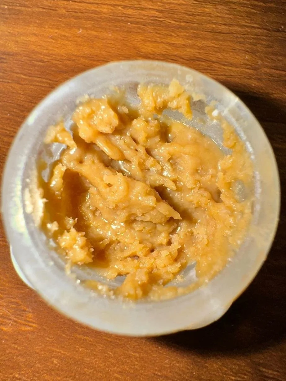 Auf diesem Bild ist eine kleine, transparente Dose mit einer gelb-orangefarbenen Paste zu sehen, die likely Erdnussbutter ist, auf einem Holztisch.