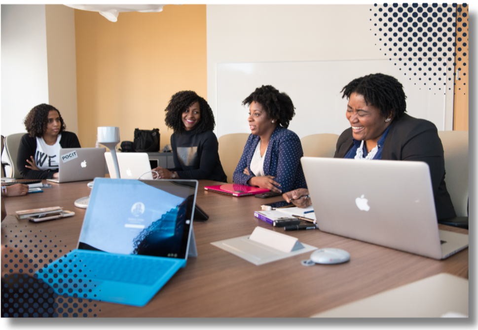 Four women with curly hair sitting at a conference table with laptops, engaging in a meeting or discussion.