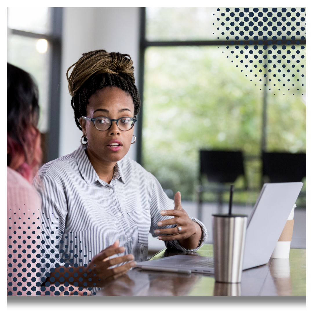 A woman with glasses and dreadlocks sitting at a table, engaged in conversation, with a laptop, tumbler, and a hand gesturing.