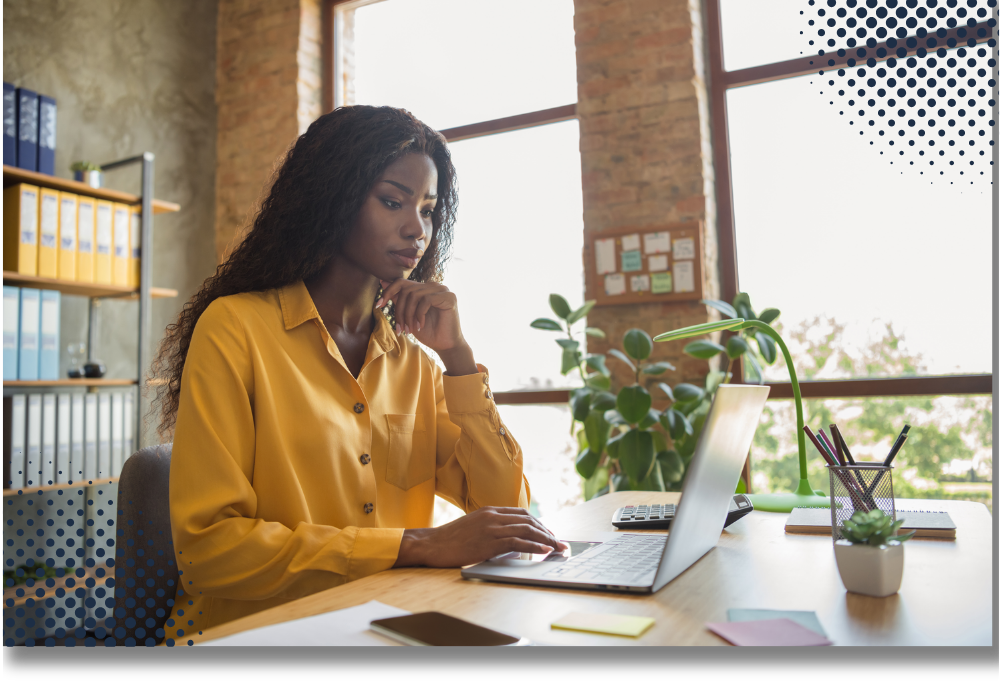 A woman in a yellow blouse working on a laptop in a bright office with large windows, a green plant, and office supplies on the desk.