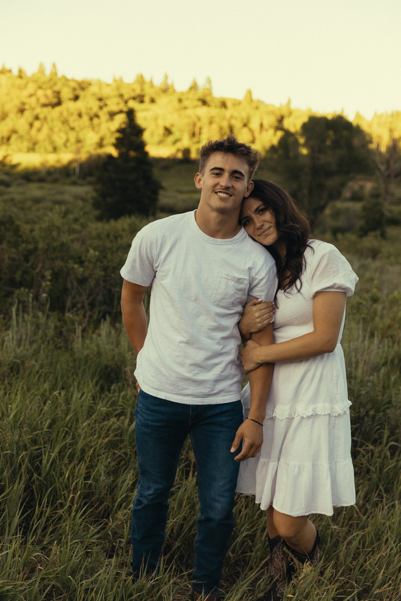 A young man and woman standing close together outdoors in a grassy field, with trees and a hill in the background, during the late afternoon or early evening.