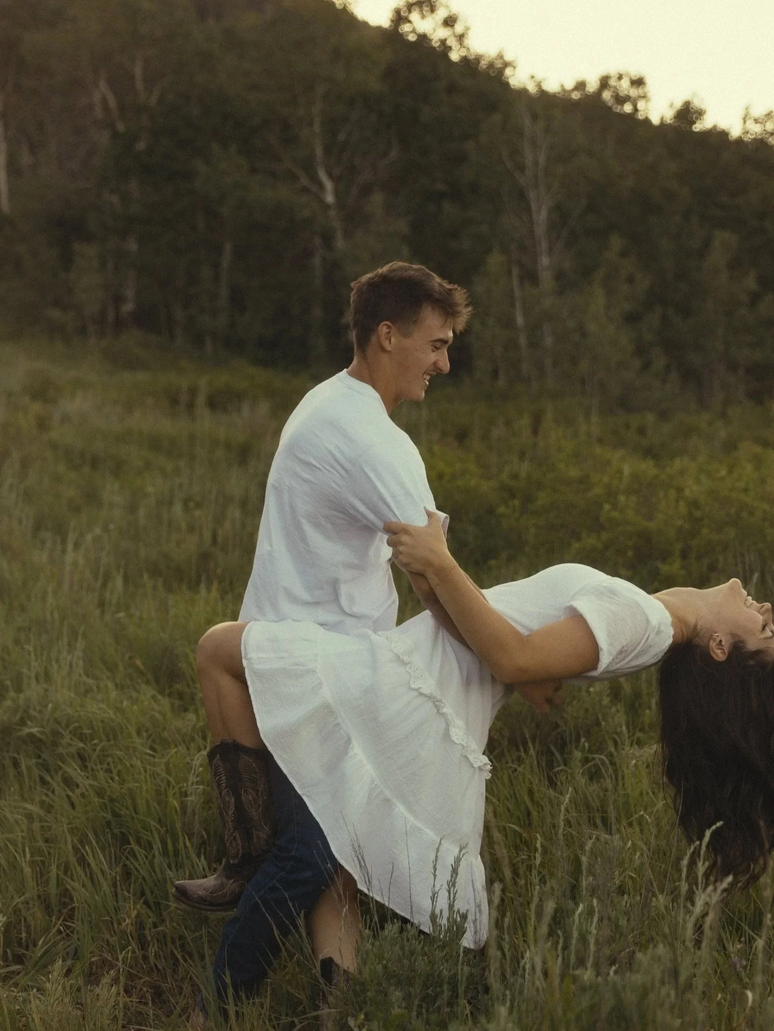 Two people, a man and a woman, engaging in a dance or playful pose outdoors in a field with tall grass, surrounded by trees, during what appears to be sunset or early evening.