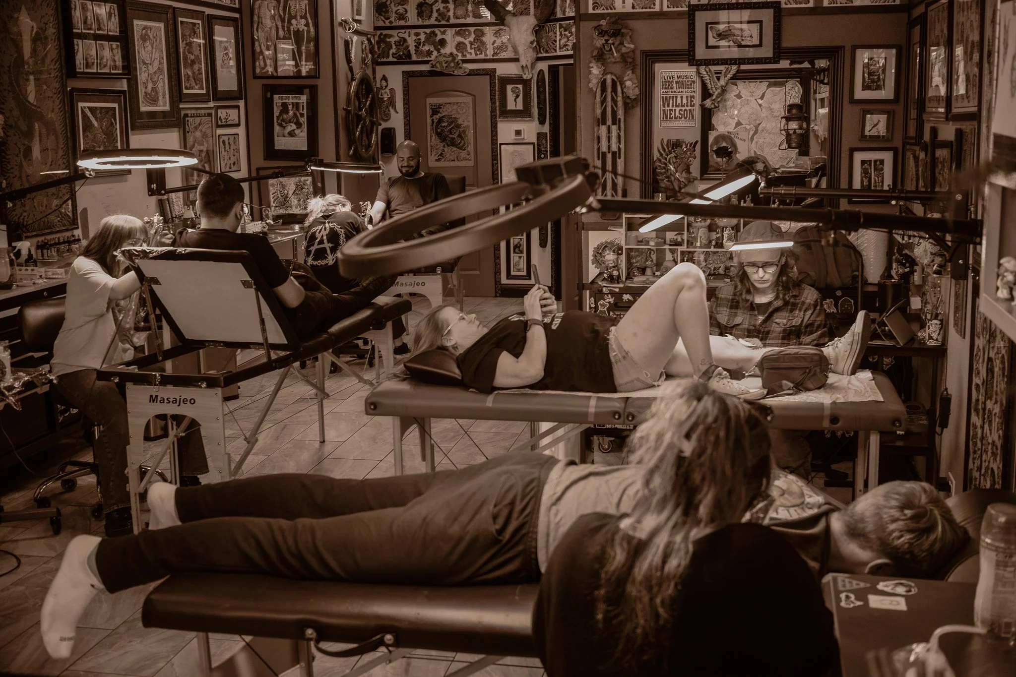 Tattoo studio with people receiving tattoos, artwork, decorations, and posters on walls, and tattoo equipment visible.