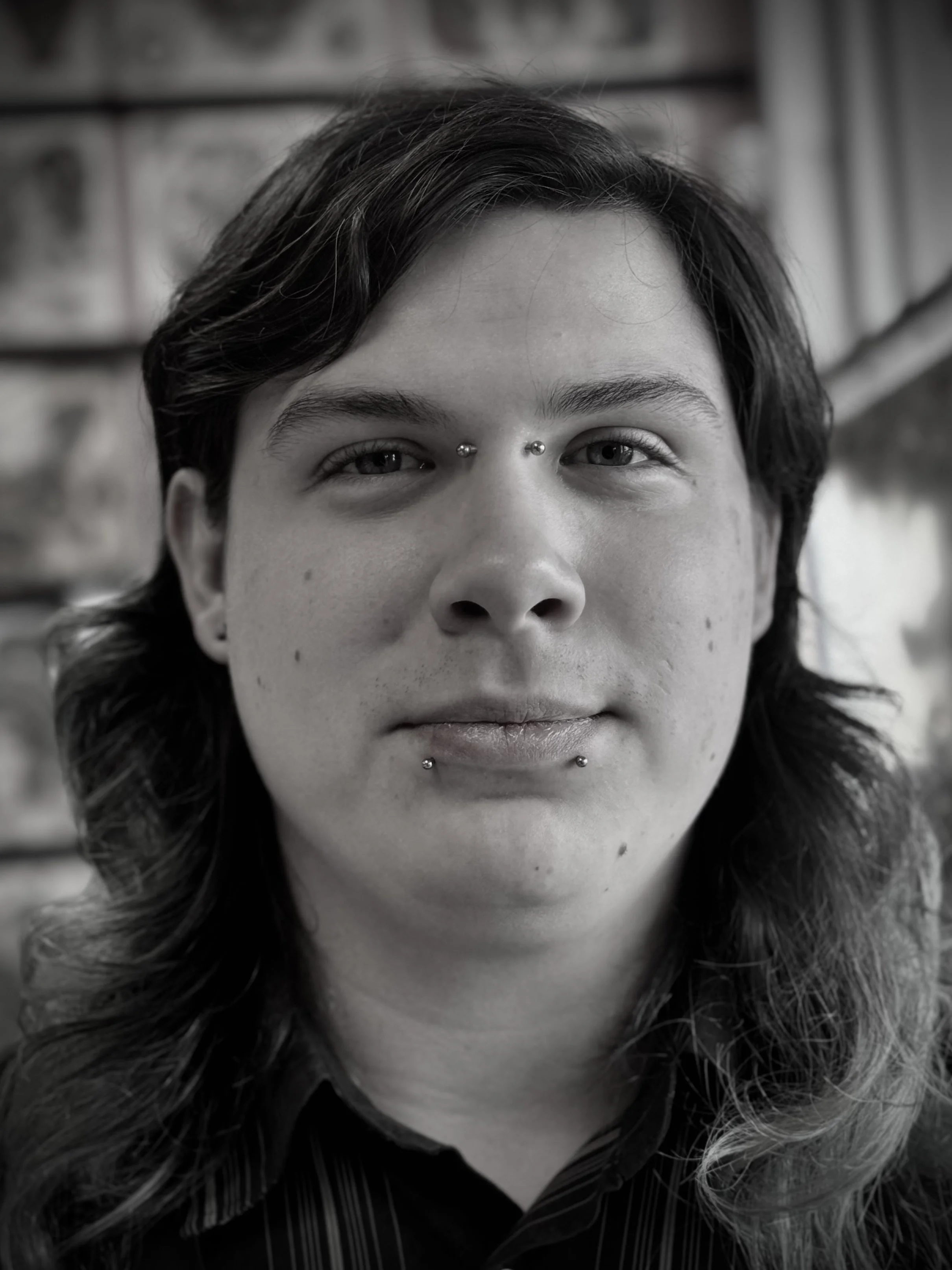 Black and white close-up of a person's face with dark, wavy hair and facial piercings including two studs on the eyebrows and two below the lower lip.