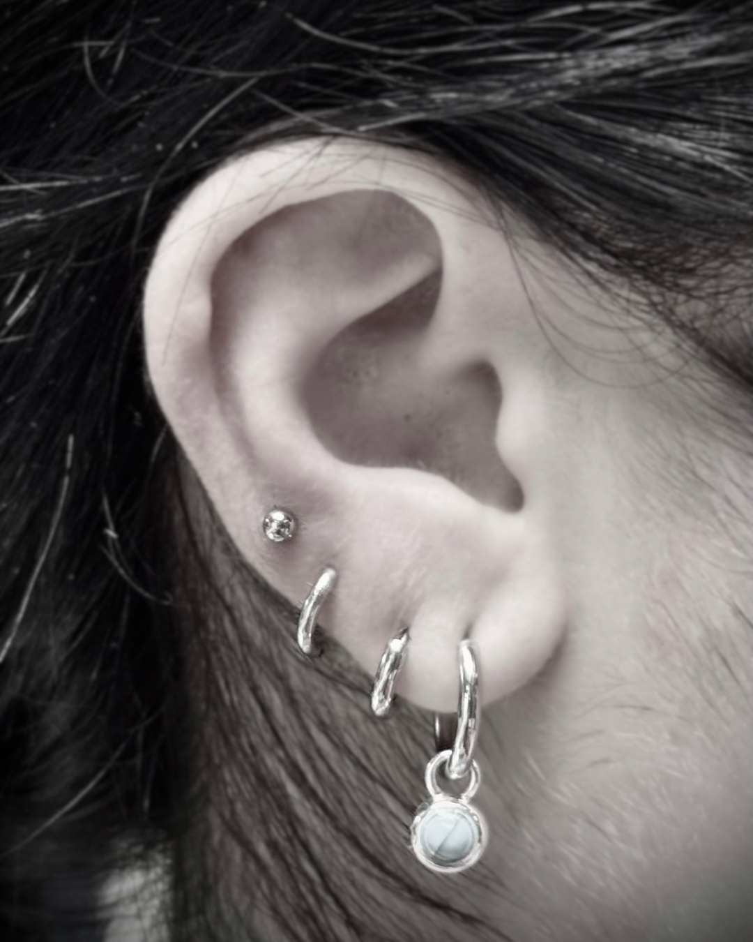 Close-up of a human ear with multiple silver earrings, including a small diamond stud and a dangling earring with a circular stone, along with dark hair partially covering the ear.