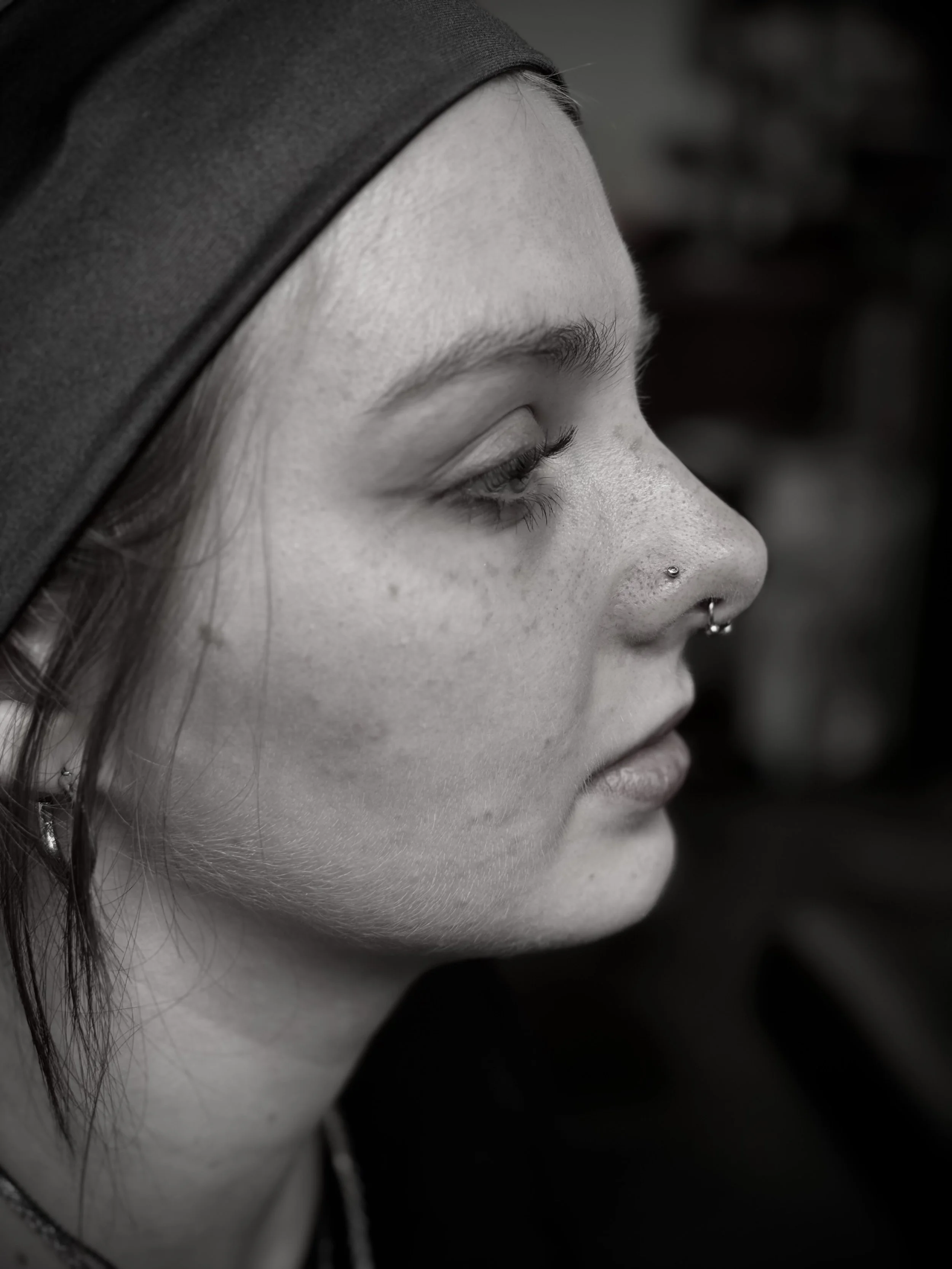 Close-up black and white photo of a woman's face in profile with her eyes closed, wearing a head covering and a septum piercing.