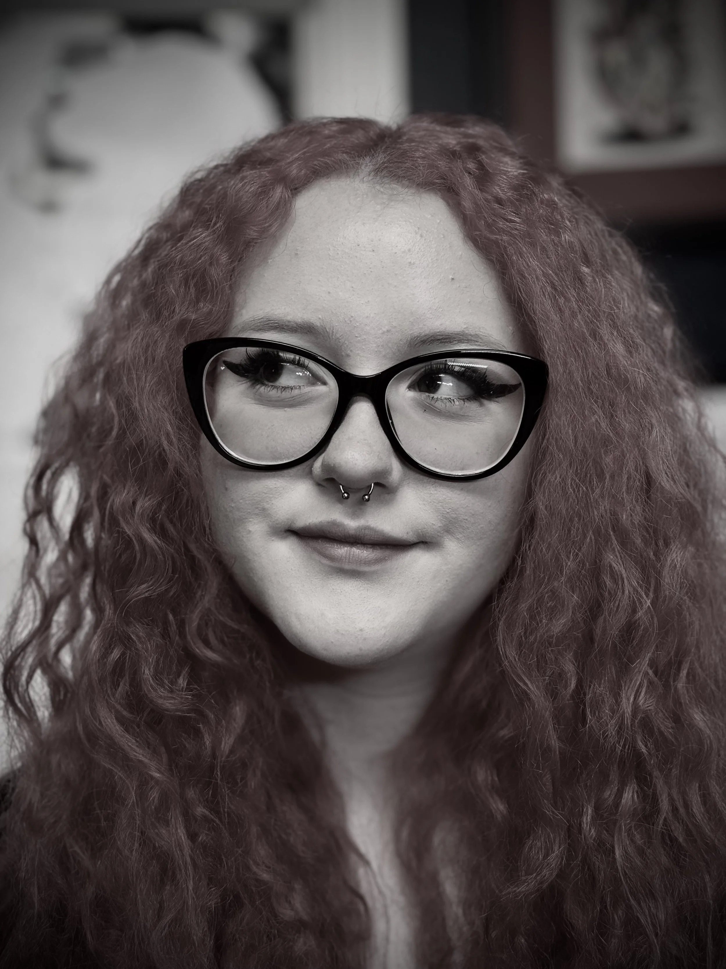 Close-up of a woman with curly red hair, wearing large black glasses and a nose ring, smiling slightly.