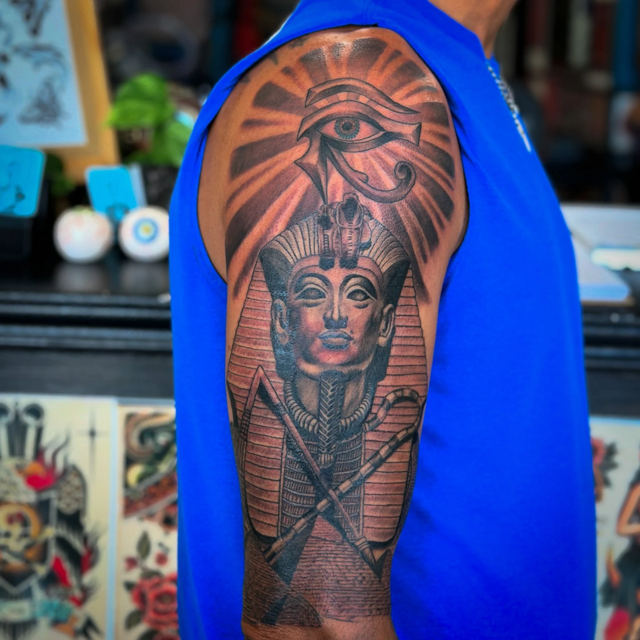 Tattoo of the Eye of Horus on a person's upper arm with sun rays background and a sphinx below, dark ink with orange shading, wearing a blue sleeveless shirt.