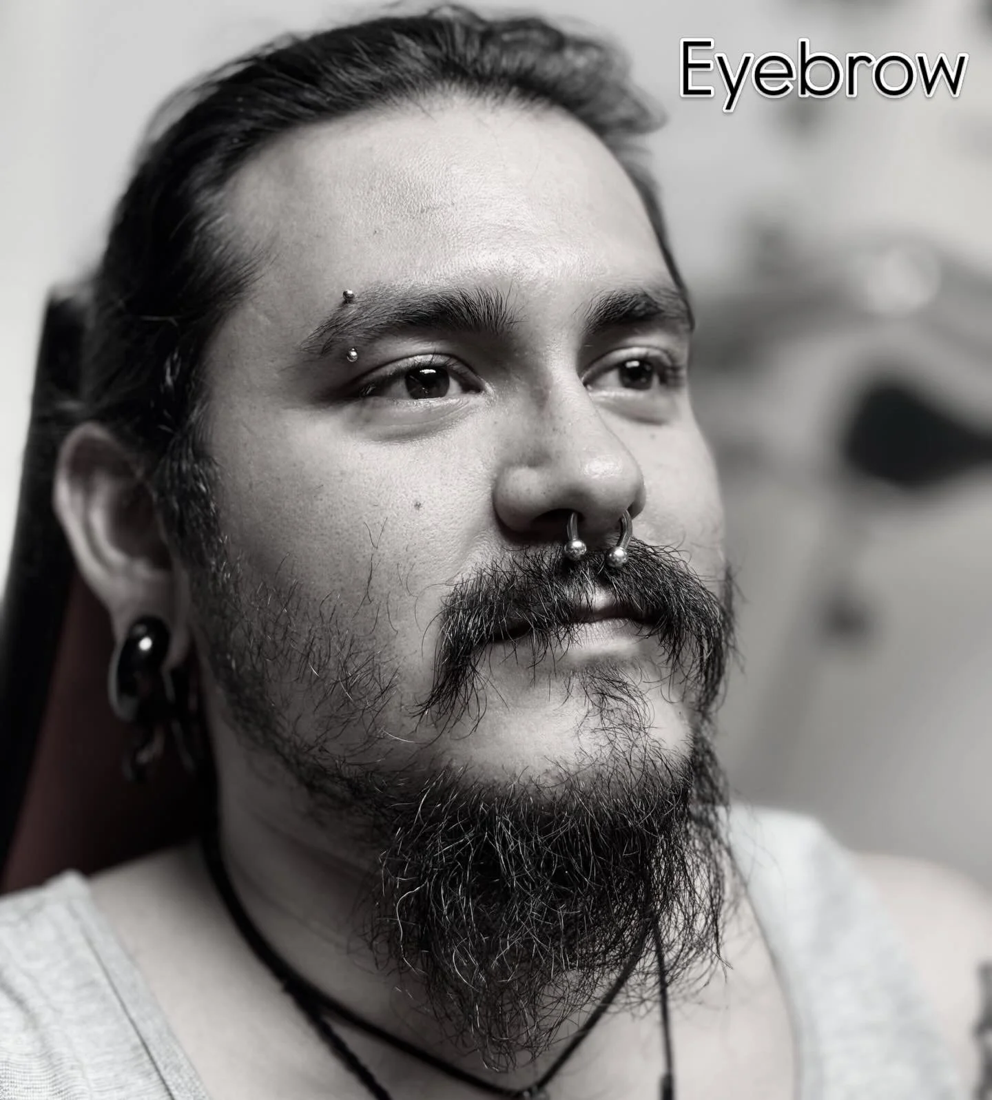 Close-up of a man with facial piercings, including a septum ring and eyebrow jewelry, with a beard and mustache, wearing a white shirt and a necklace, looking to the side, with the word 'Eyebrow' in the top right corner.