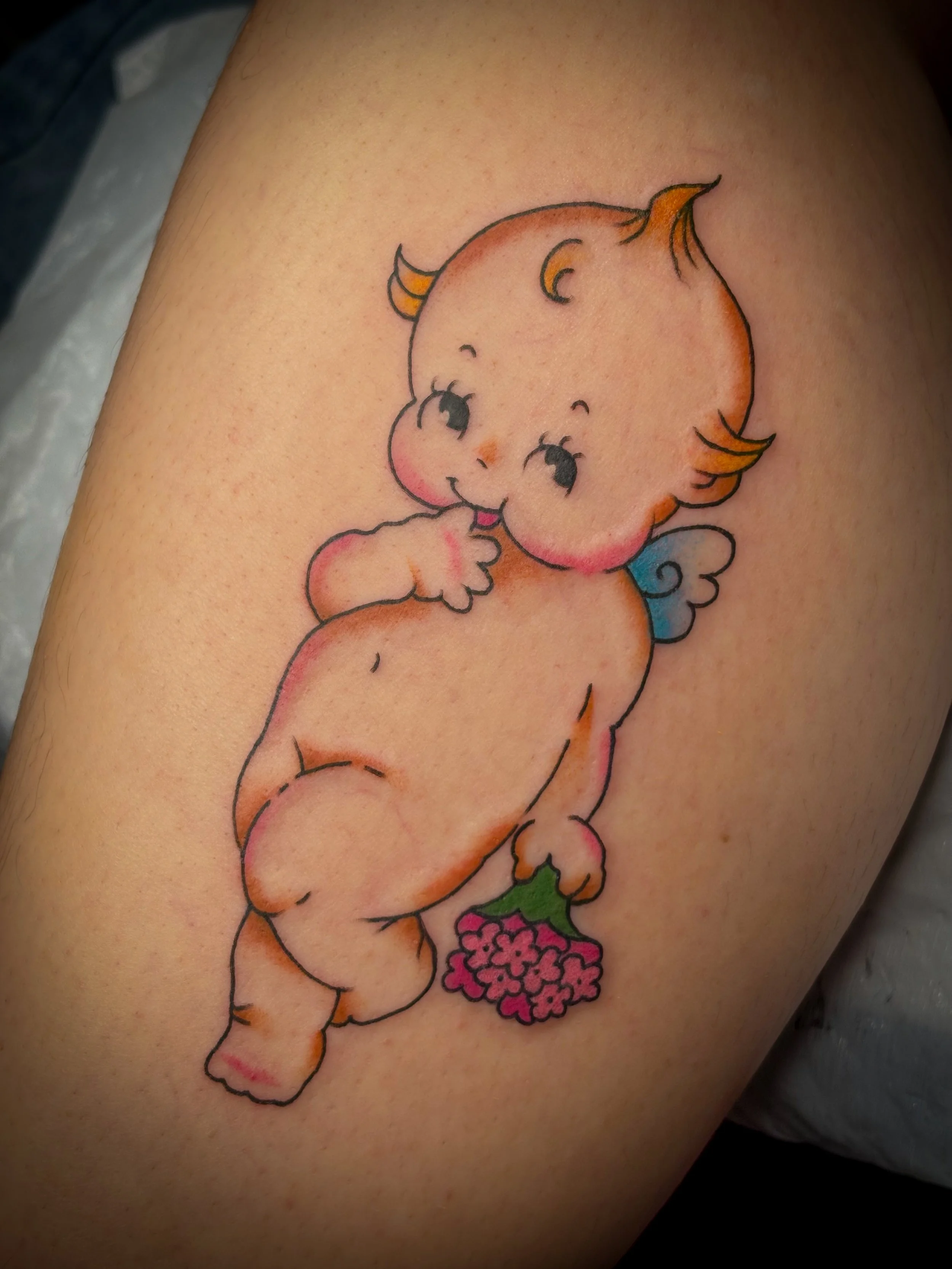 Tattoo of a quwpie. Cute, cartoon-style baby cherub with orange hair, cheek blush, and tiny wings, holding pink flowers.