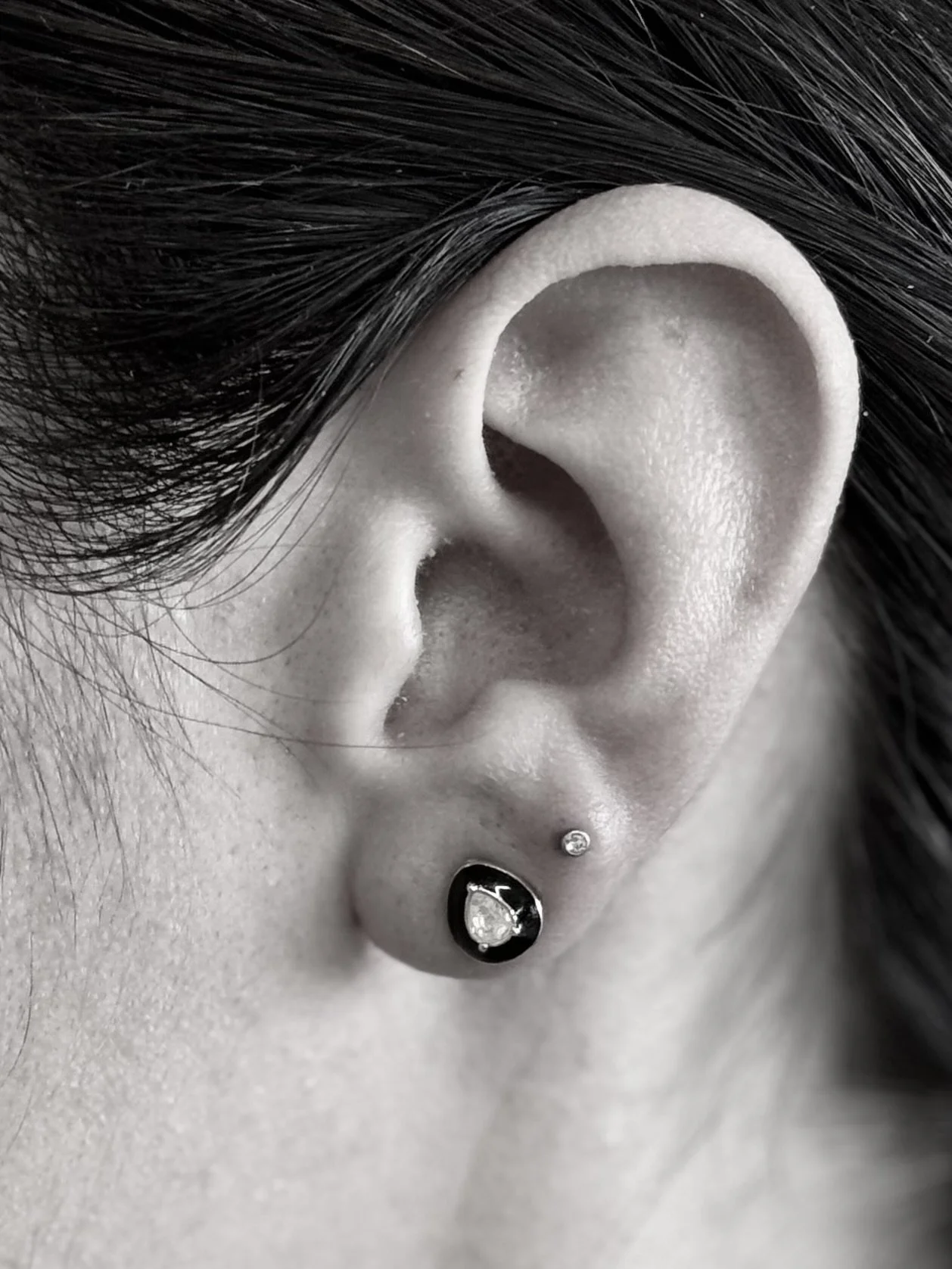 Close-up of a person's ear with multiple piercings, including a black earring with a clear gemstone, a small stud above, and a helix piercing near the top, accompanied by dark hair.
