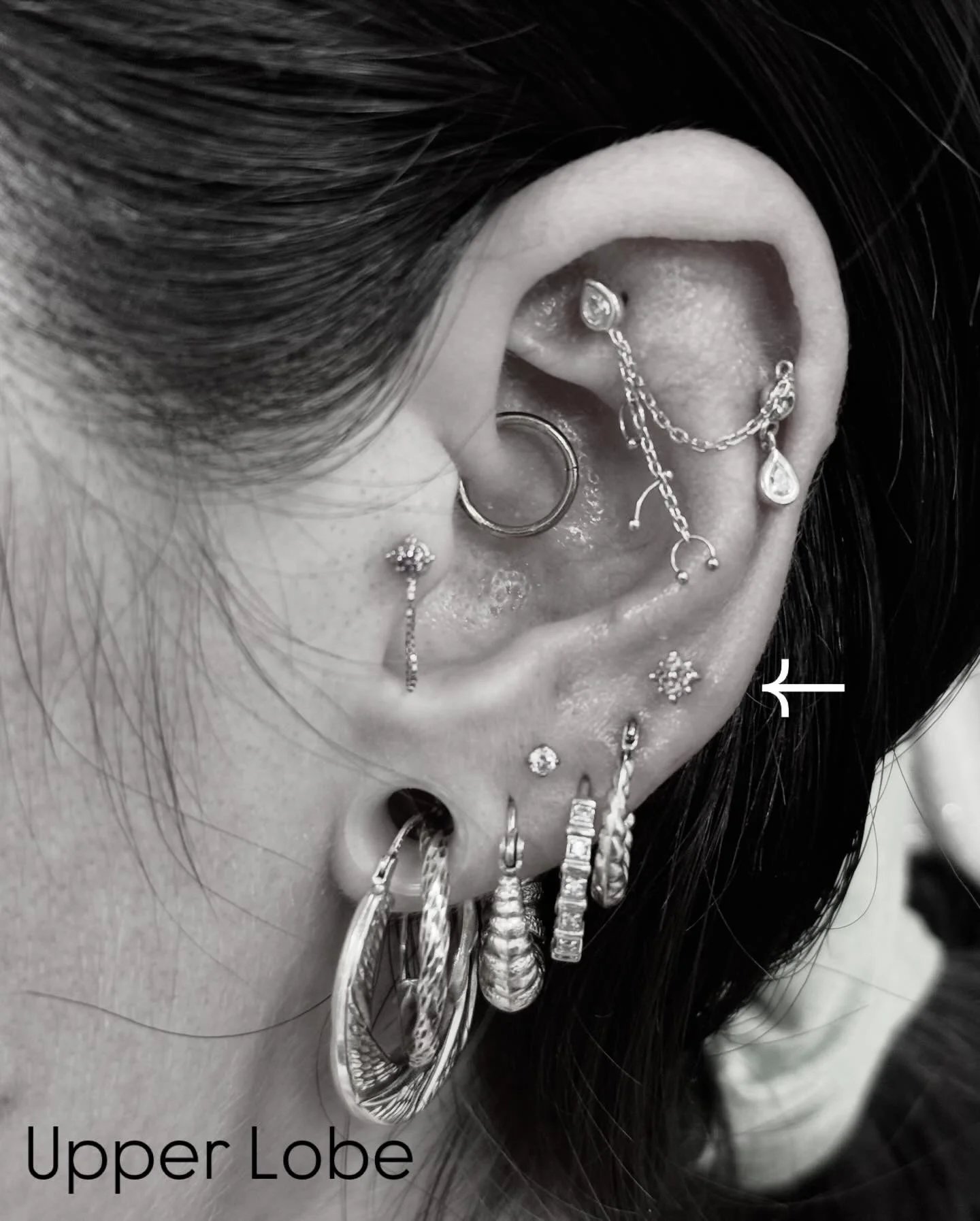 Close-up black and white photo of a woman's ear in the upper lobe area, showcasing various earrings, piercings, and jewelry, with a timestamp label pointing to the upper lobe.