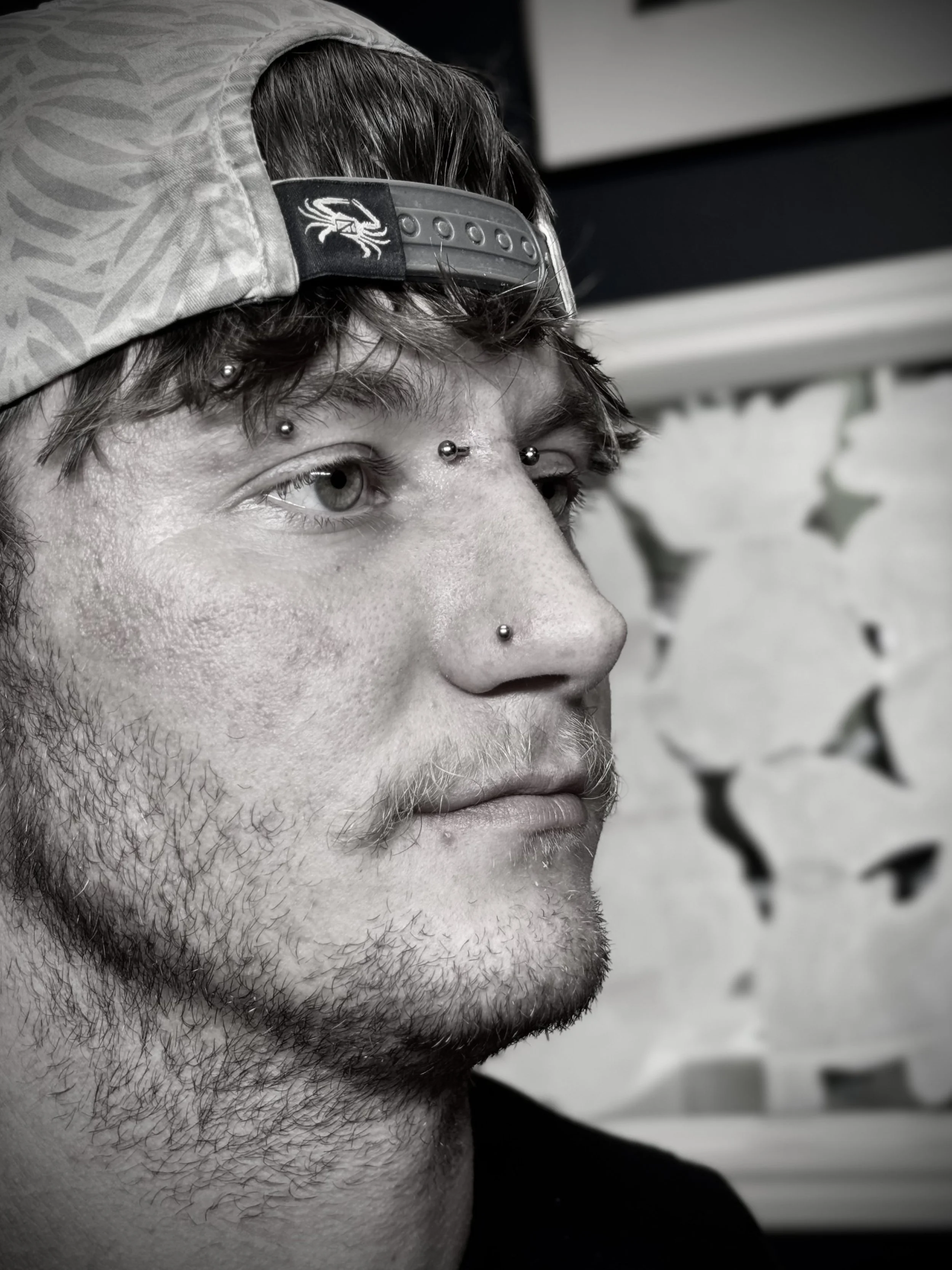 Close-up of a man's face in black and white, showing his eye, nose pierced with a stud, and facial hair. He wears a cap backward with a scorpion logo. The background includes a blurred pattern with star shapes.