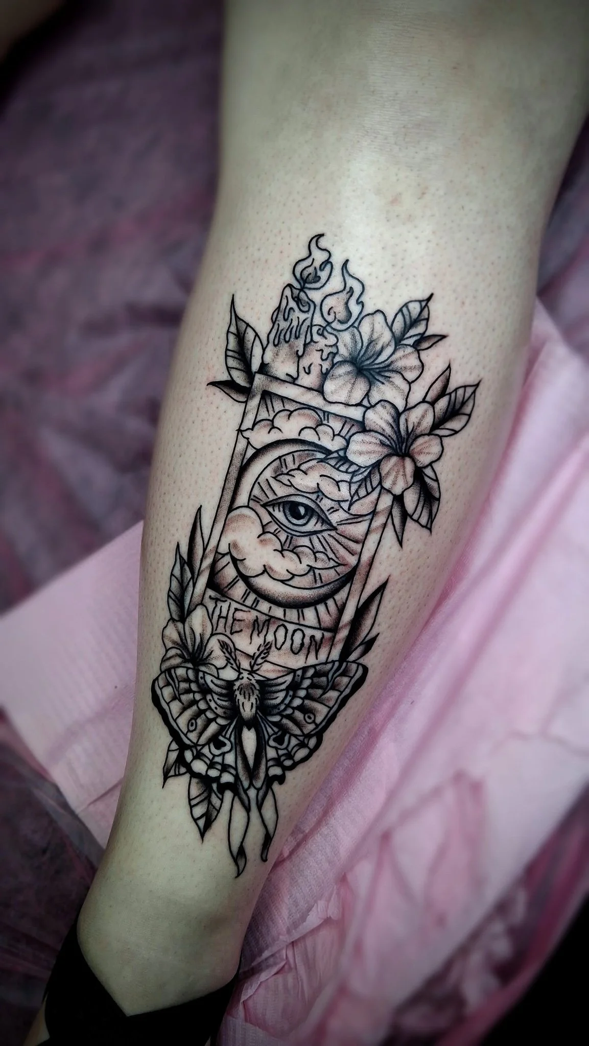 Tattoo of an eye within a moon shape, surrounded by flowers, leaves, a moth, a candle, and the phrase "The Moon" in a decorative style.