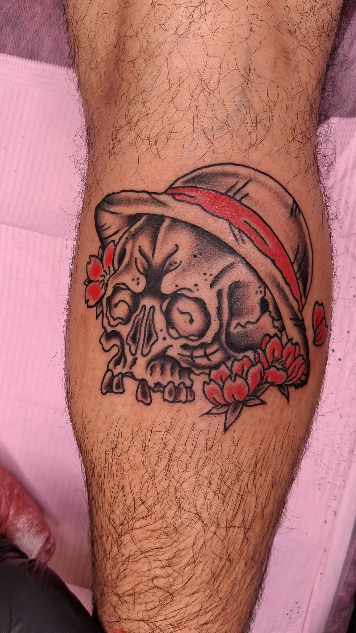 Tattoo of a skull with a red hat and flower embellishments on a person's leg.