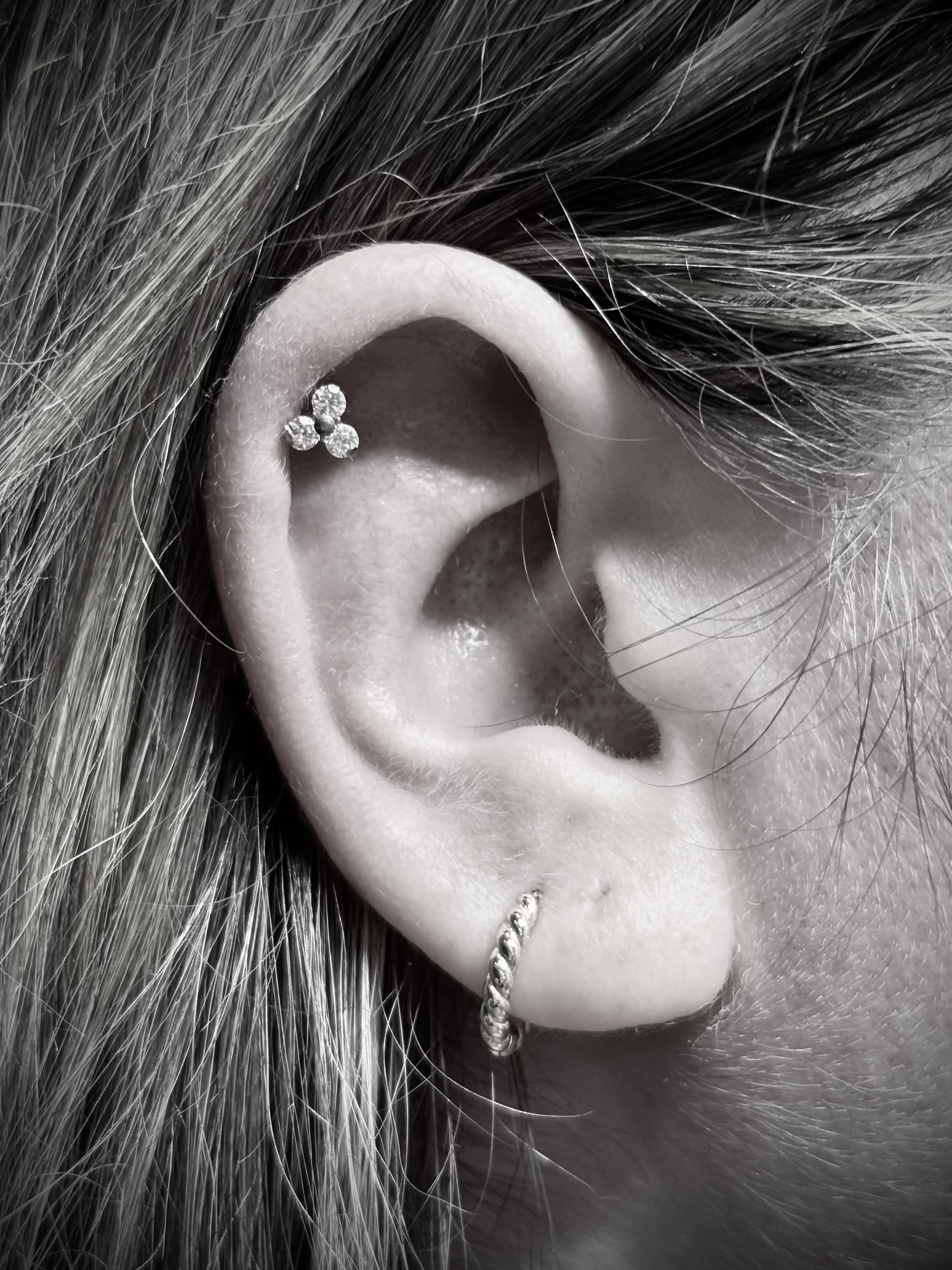 Close-up of a person's ear with multiple earrings, including a small hoop and a diamond-studded flower-shaped stud, surrounded by long hair strands.