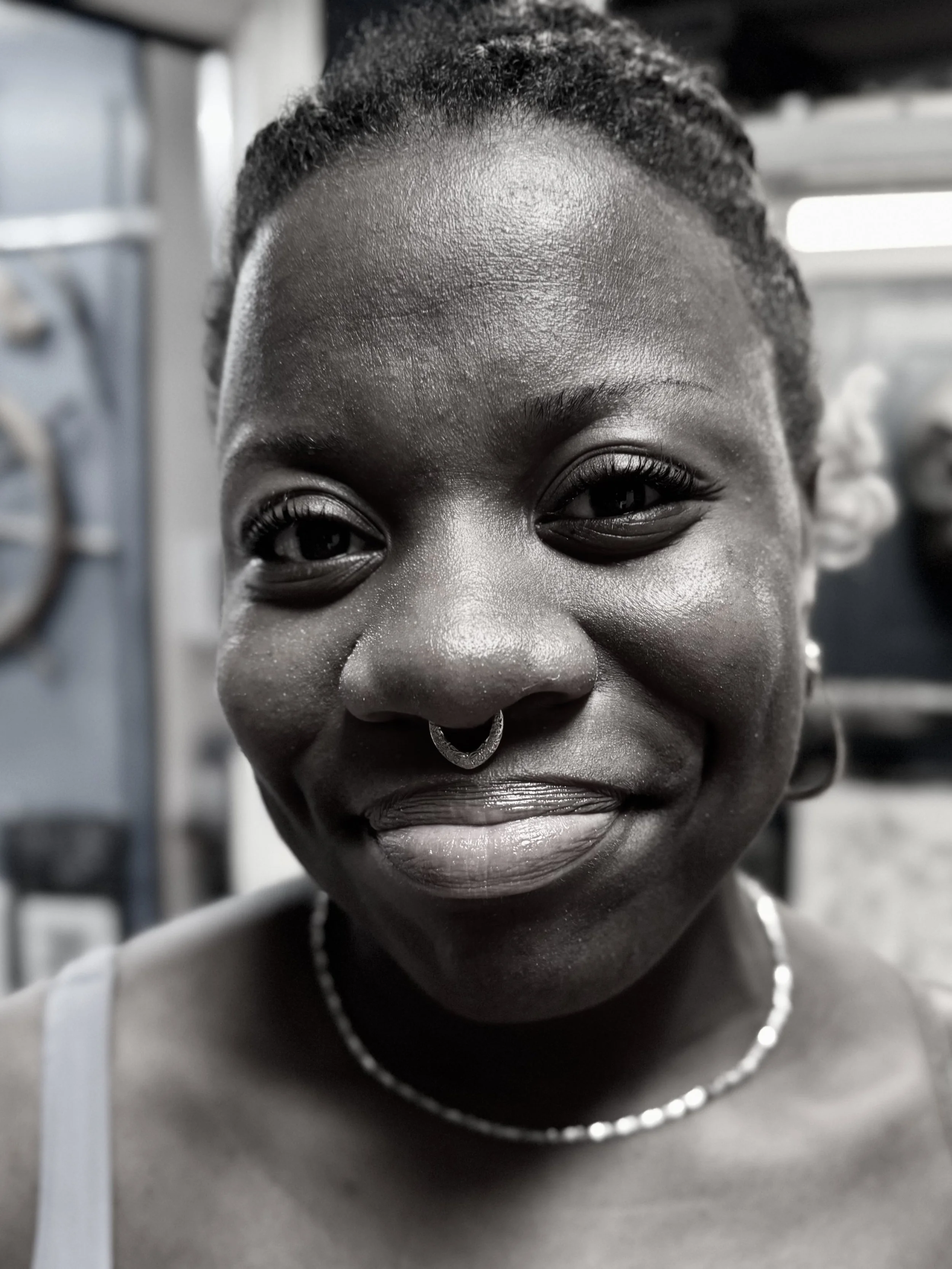 Close-up of a smiling Black woman with braided hair, wearing a ring nose and a pearl necklace, indoors with a blurred background.