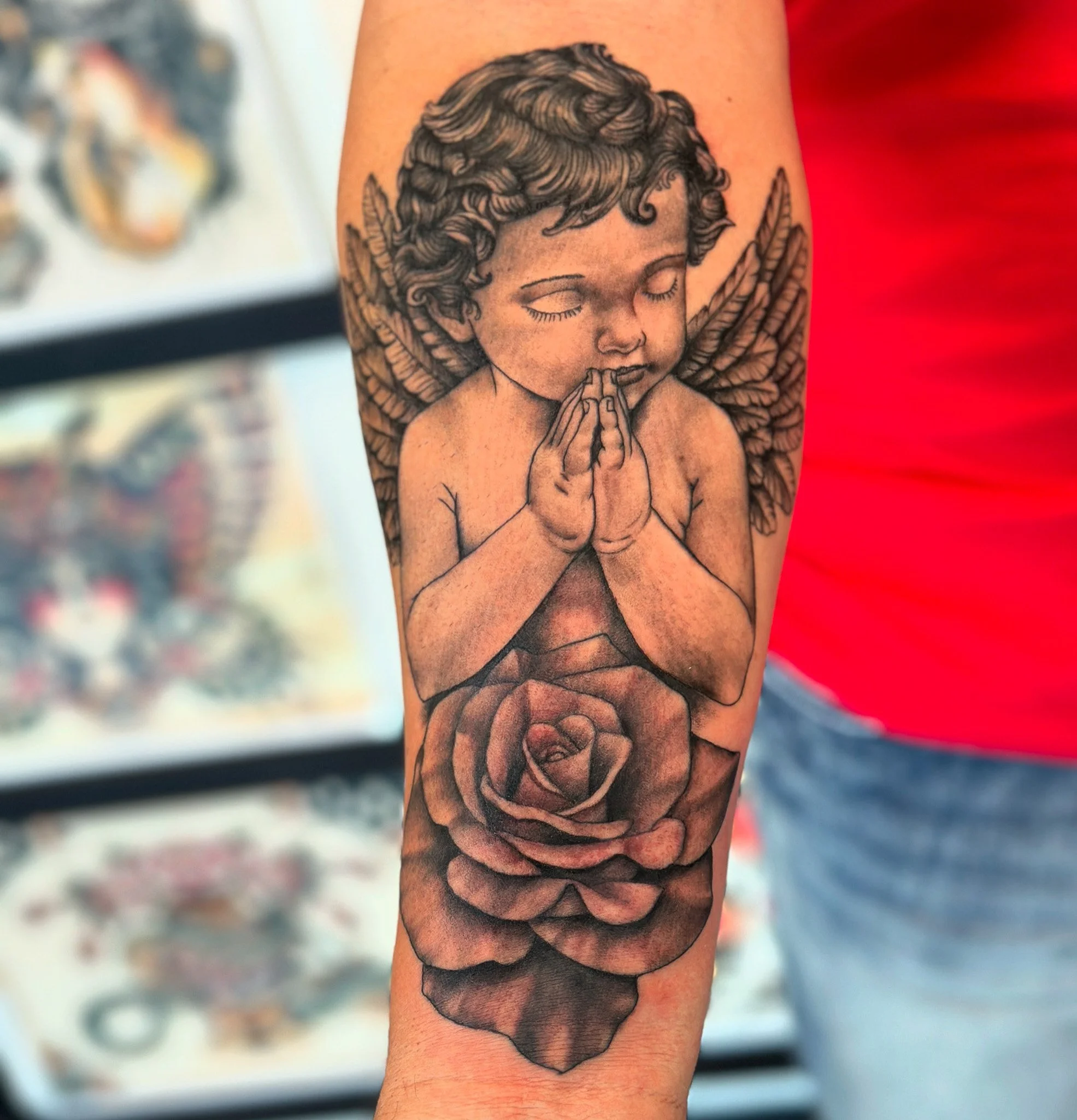 Tattoo of praying angel with closed eyes and hands, surrounded by roses, on a person's arm.