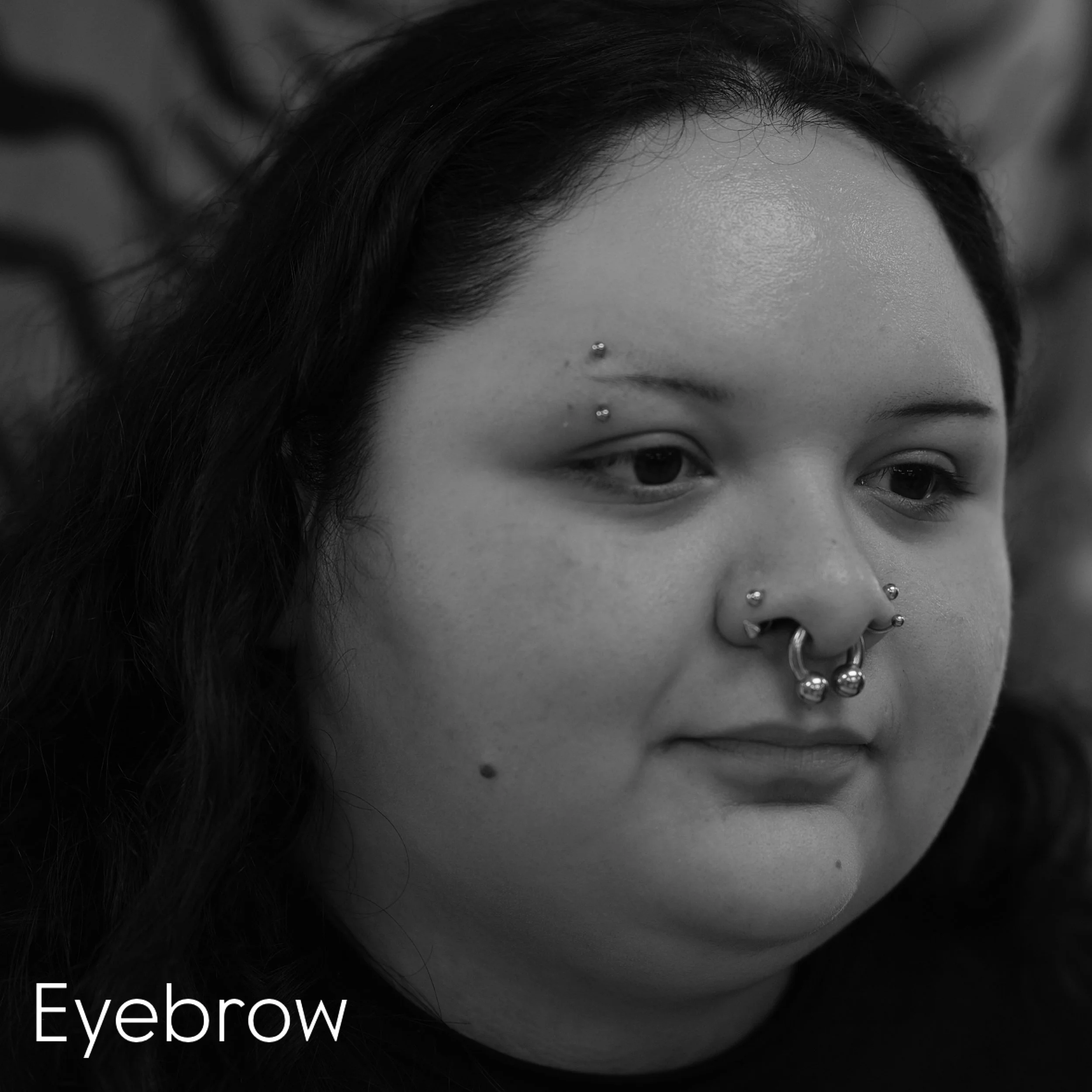 A close-up black and white photograph of a person's forehead and eye area with a facial piercing. There are large hoop earrings and the partial text 'Eyebrow' visible at the bottom.