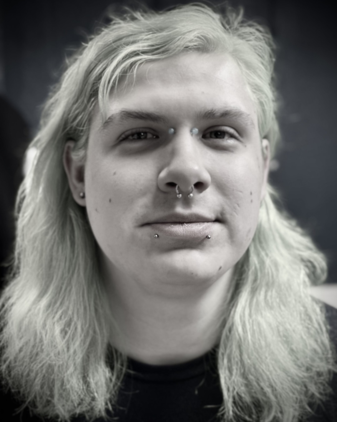 A young person with long blonde hair and multiple facial piercings, including a septum piercing, labret piercings, and eyebrow piercings, looking directly at the camera with a slight smile.