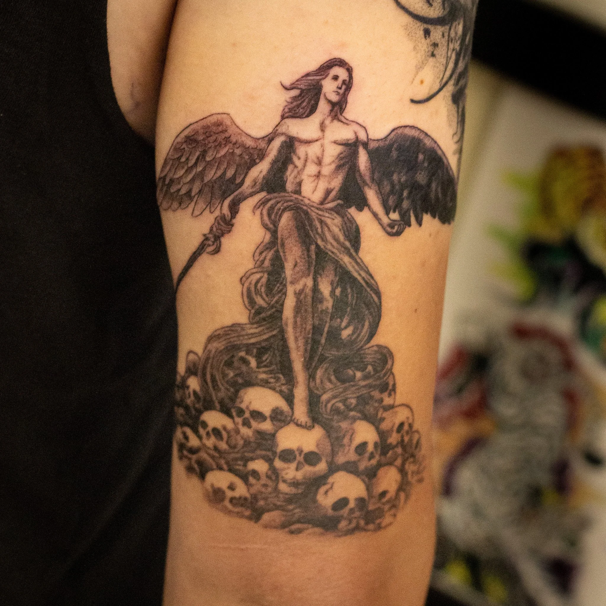 Tattoo of a winged with flowing hair, sitting in a relaxed pose, on a person's upper arm.