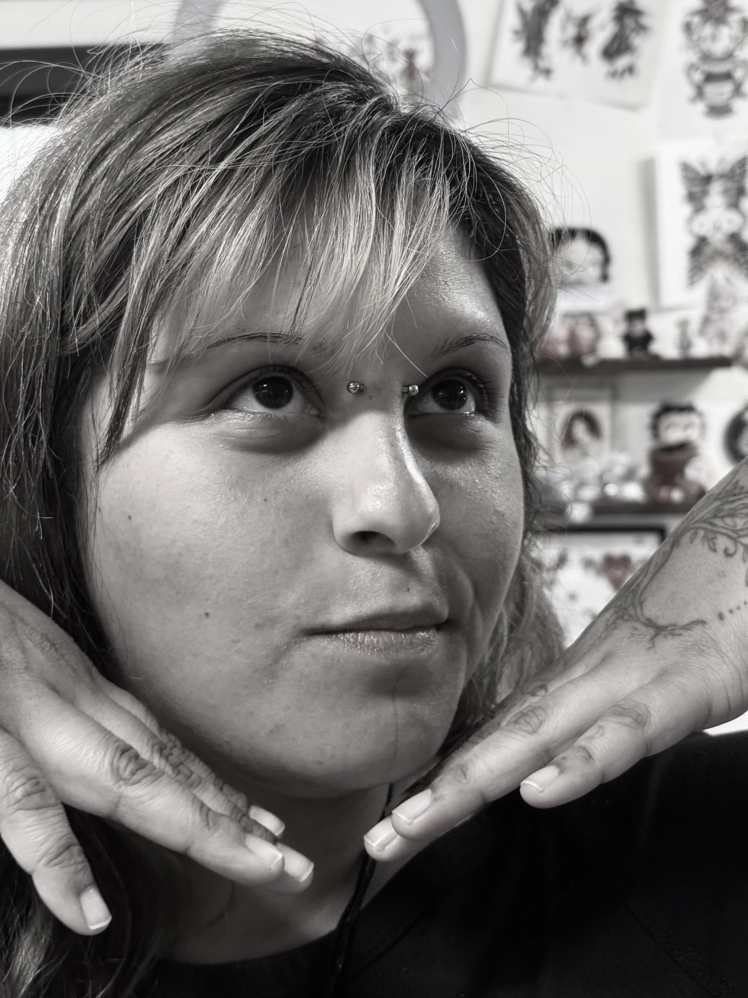 Close-up of a woman's face with dark hair, wide eyes looking upward, and hands framing her face with fingers touching her cheeks, in black and white.