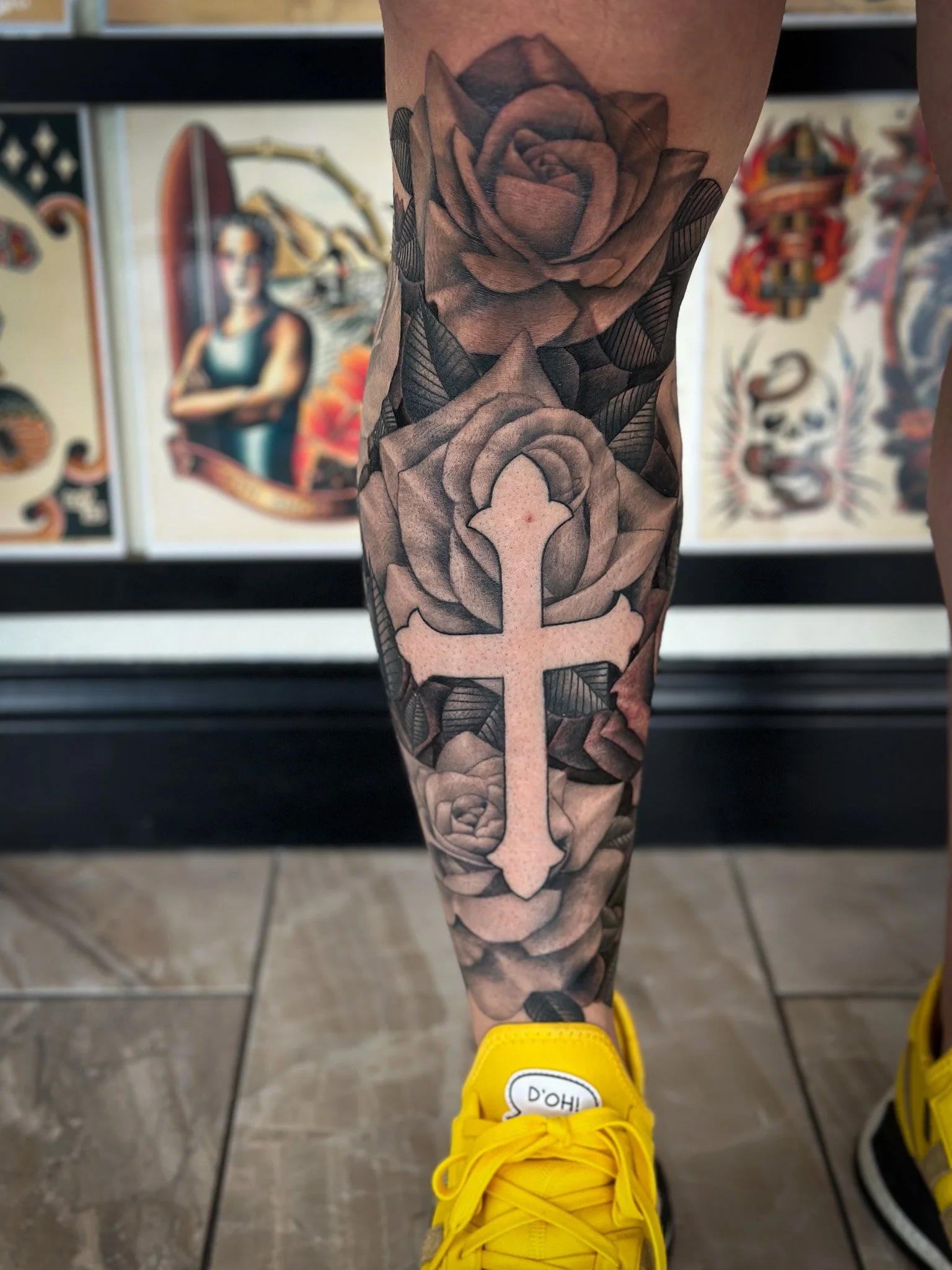 Black and gray tattoo of roses and a cross on a person's leg.