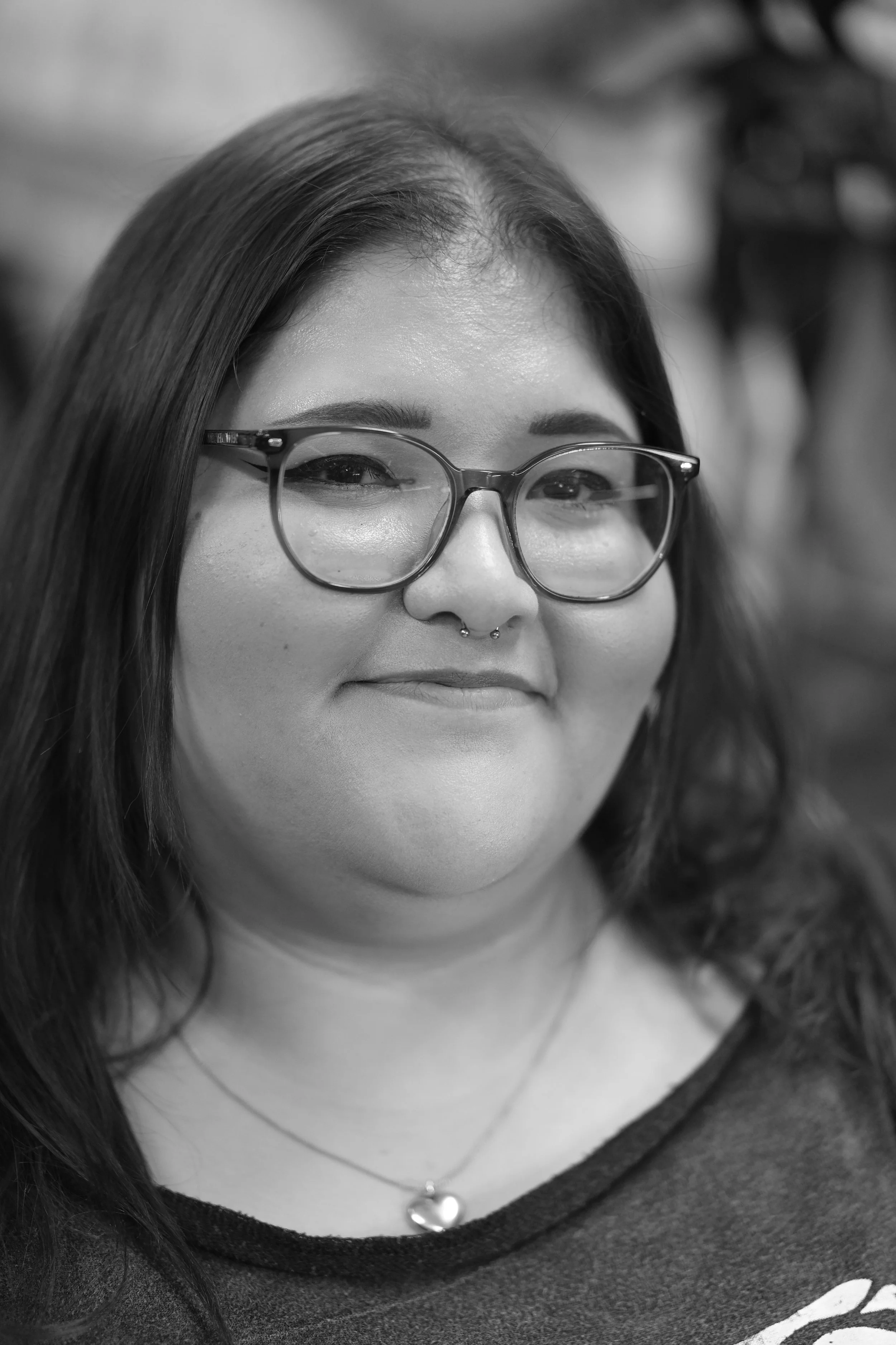 A woman with long dark hair, wearing glasses and a heart-shaped necklace, smiling slightly.