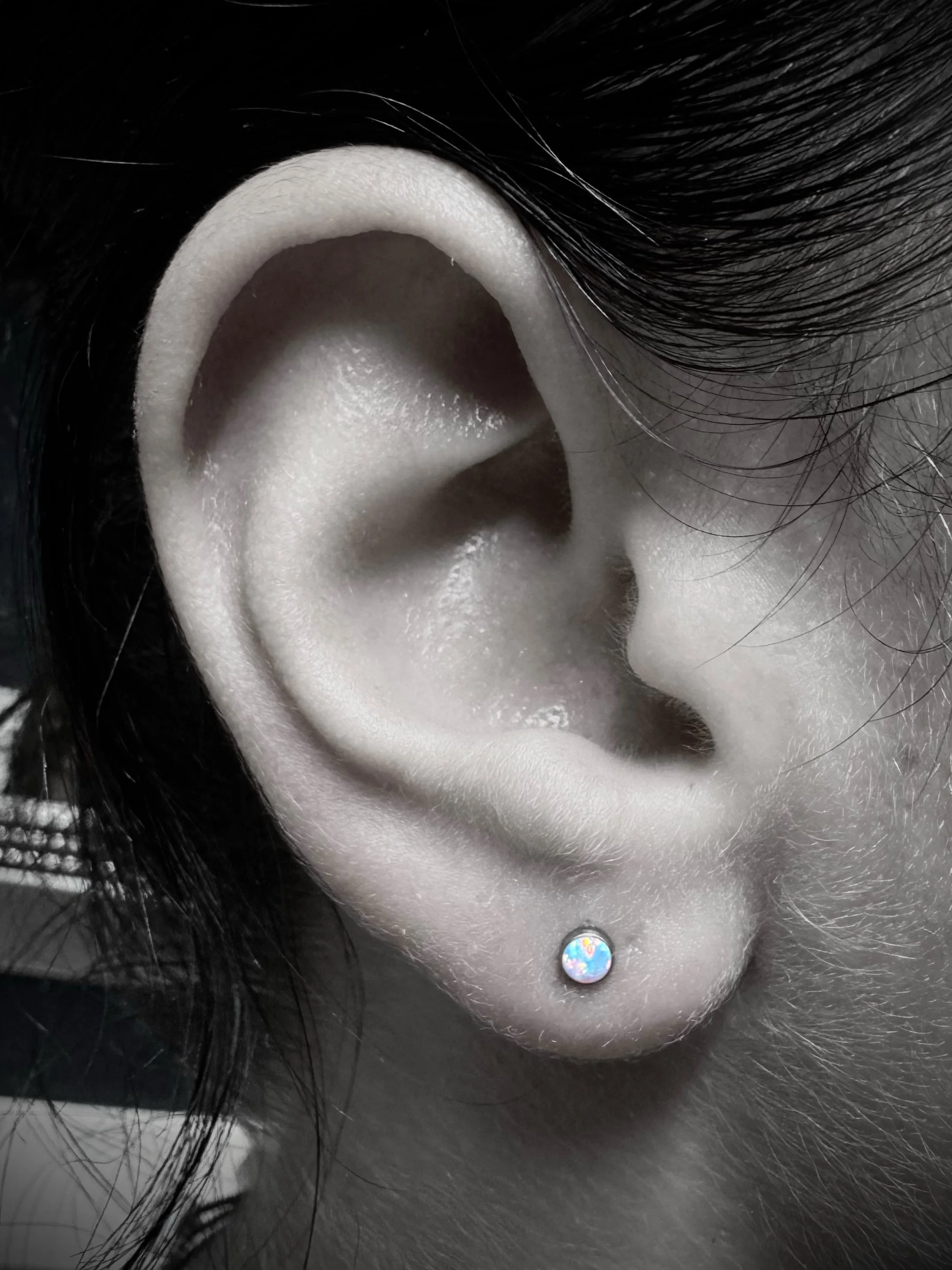 Close-up of a human ear with black hair surrounding it, and a small colorful circular earring on the lower part of the ear.