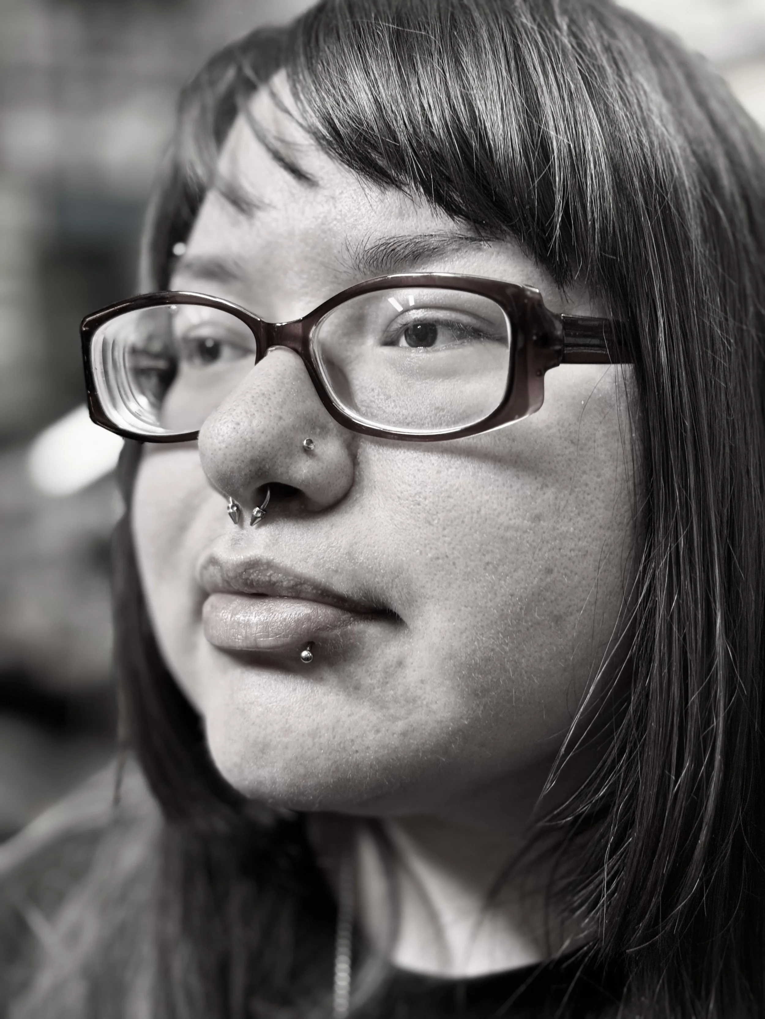 Close-up of a person with glasses, multiple facial piercings, and straight hair.