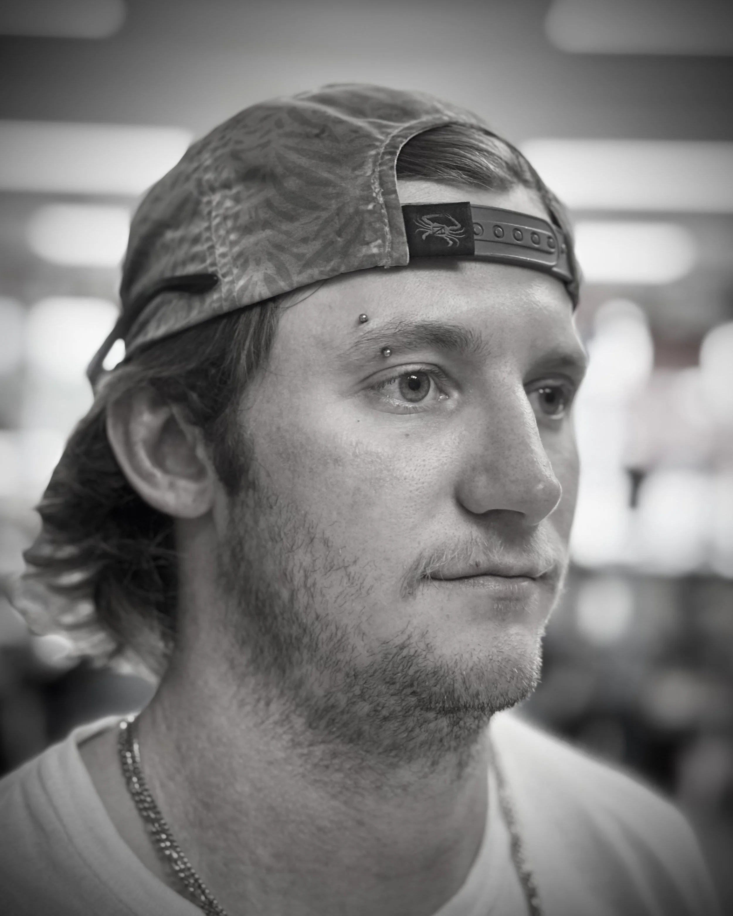 Close-up of a young man with a baseball cap, a pierced eyebrow, and facial hair, looking to the side, with a blurred background.