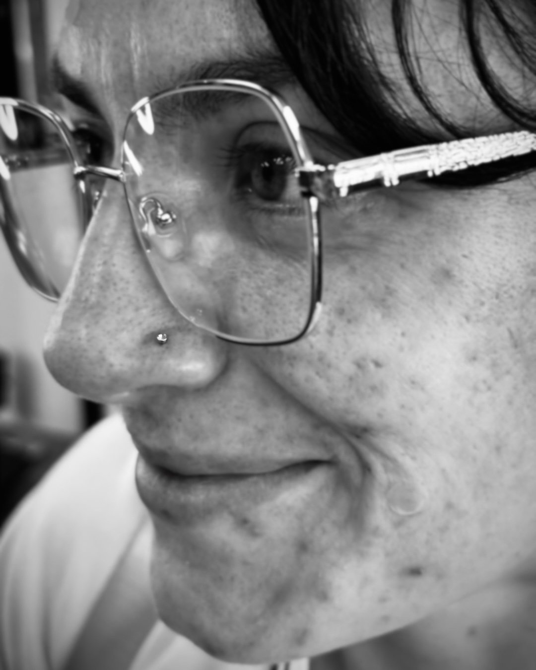 Close-up black and white photo of a woman wearing glasses with a nose piercing, smiling, and a tear rolling down her cheek.