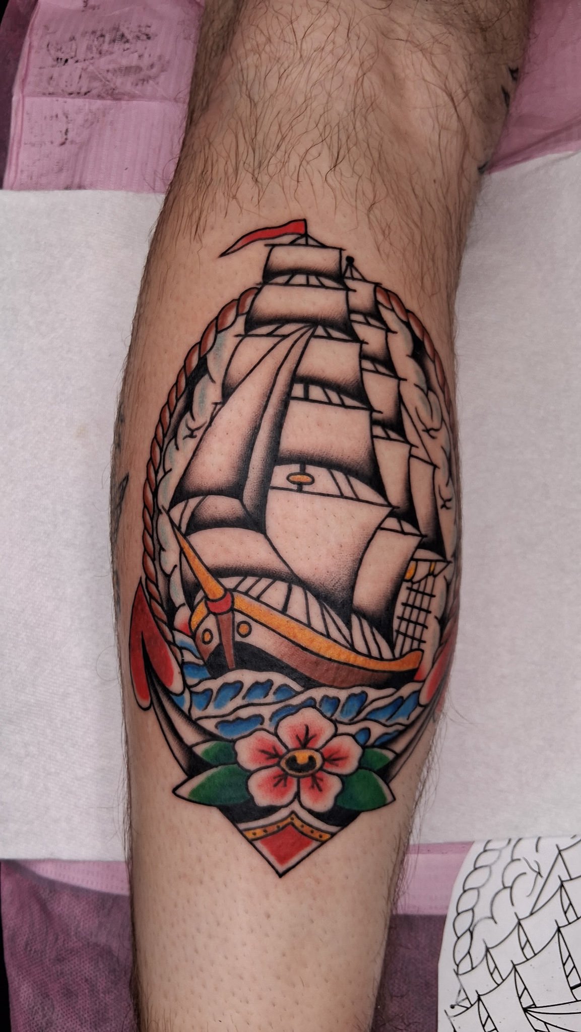 Colorful tattoo of a traditional sailing ship inside a circular design on a person's arm, surrounded by nautical elements and a flower at the bottom.
