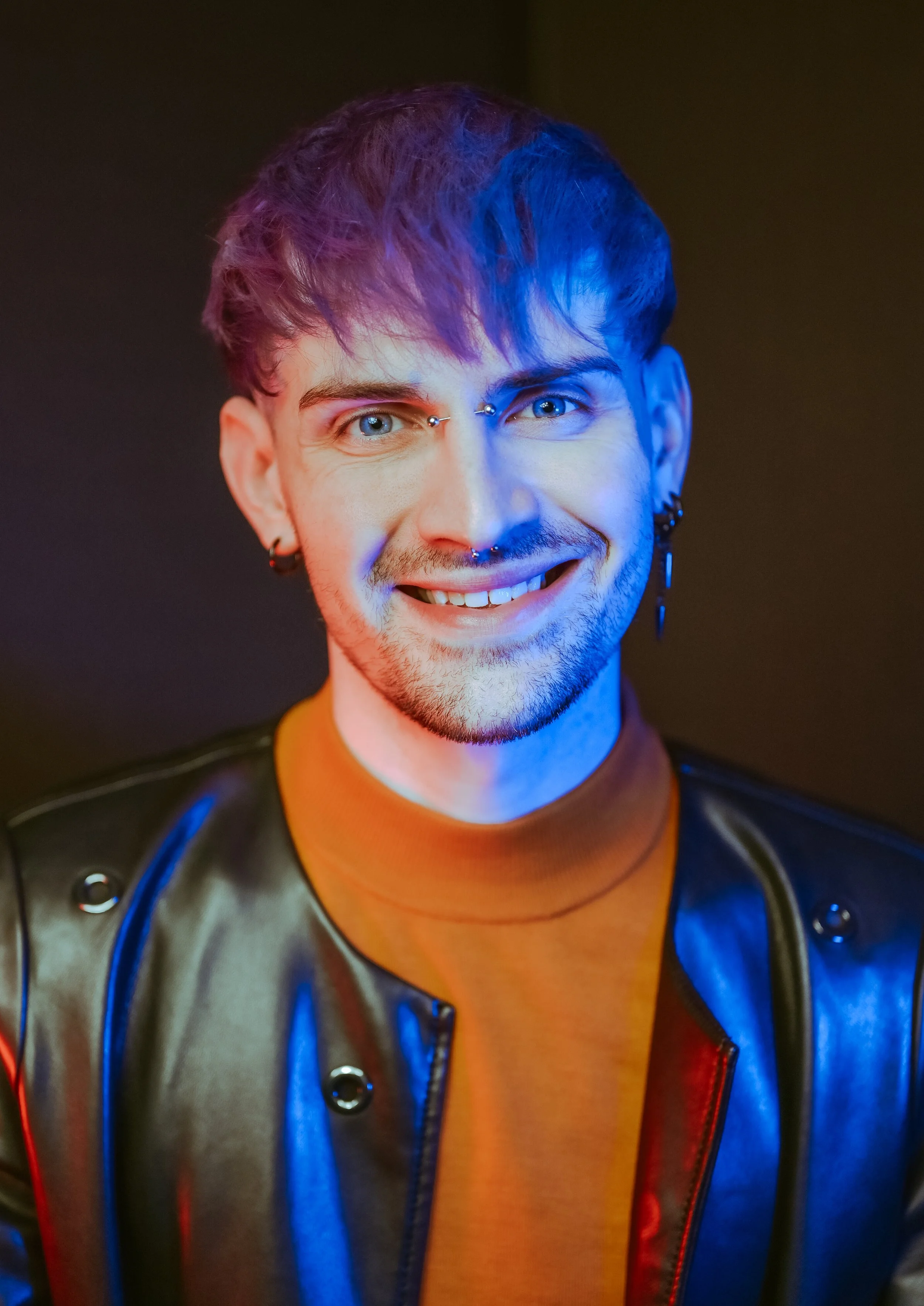 A young man with blue eyes and facial piercings smiling, wearing earrings, a septum ring, and a leather jacket, with colorful lighting on his face.