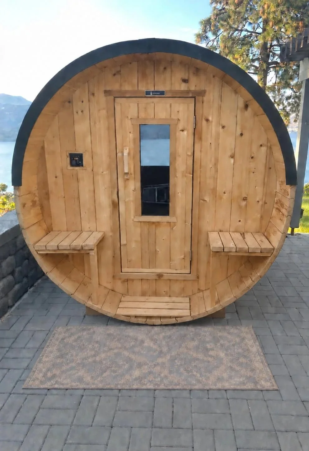 A round wooden sauna with benches on both sides, a door with a small window in the center, and a black strap handle. It is situated outdoors on a stone patio with a rug in front, with trees and mountains in the background.