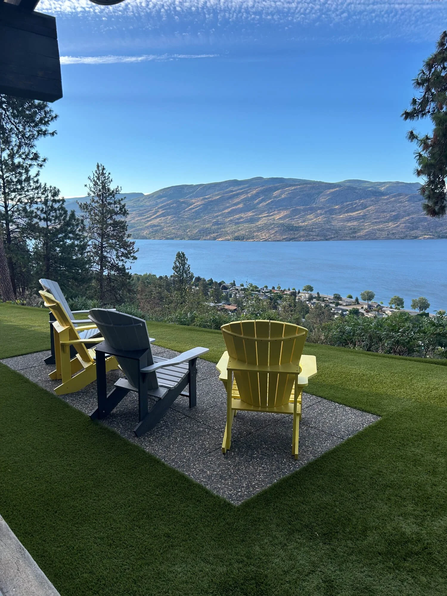 Colorful outdoor patio area with yellow and dark blue Adirondack chairs facing a lake and mountain landscape in the background. The patio has a small gravel section and well-maintained green grass.