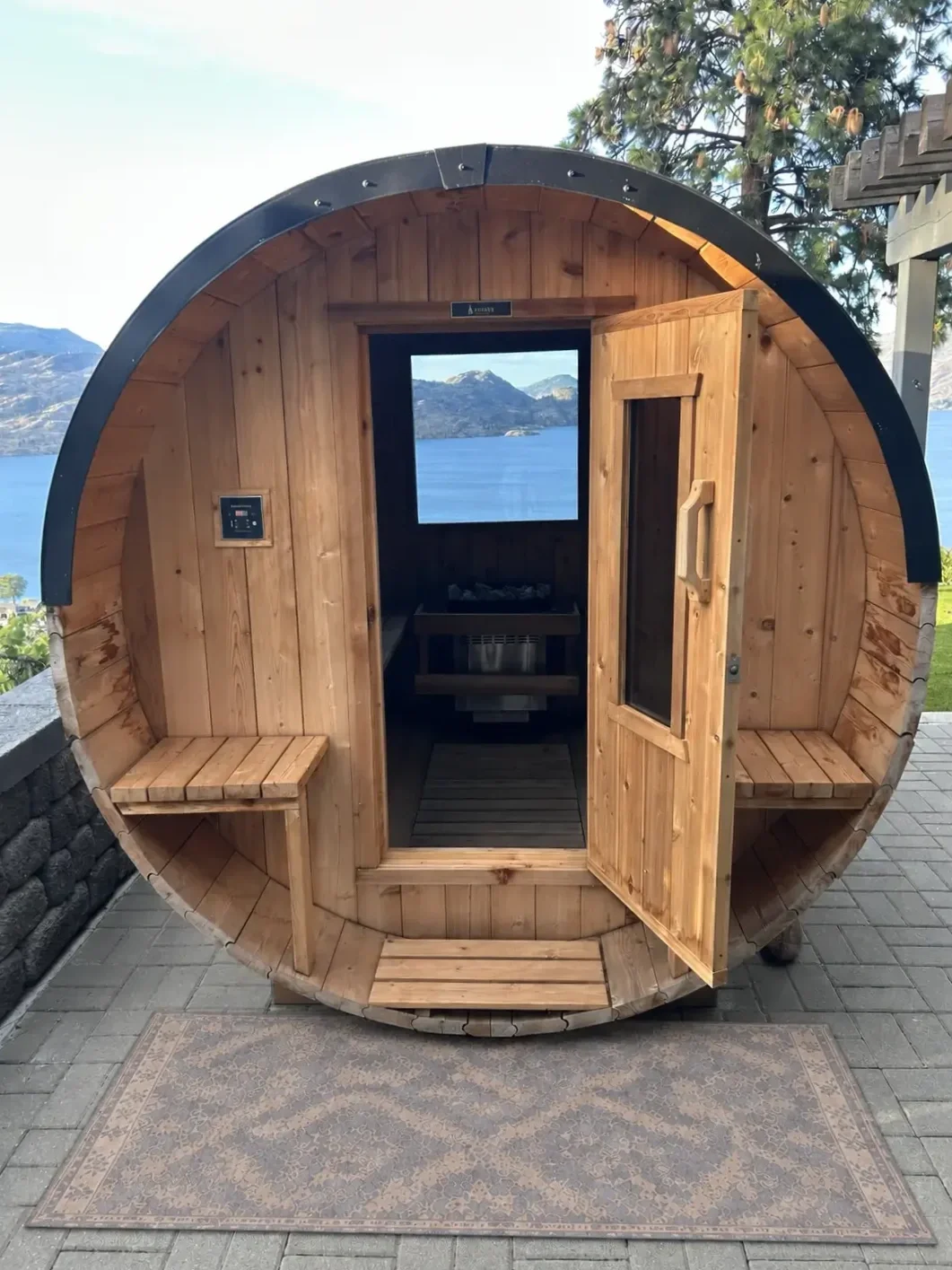 Small wooden barrel-shaped outdoor structure with benches on each side, open door revealing a sauna interior, scenic mountain and lake view in the background.