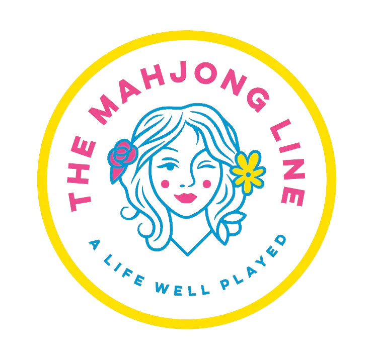 Logo for The Mahjong Line, featuring a stylized woman with long wavy hair, pink cheeks, a pink rose and a yellow flower in her hair, surrounded by pink and blue text on a black background, with a yellow circular border.