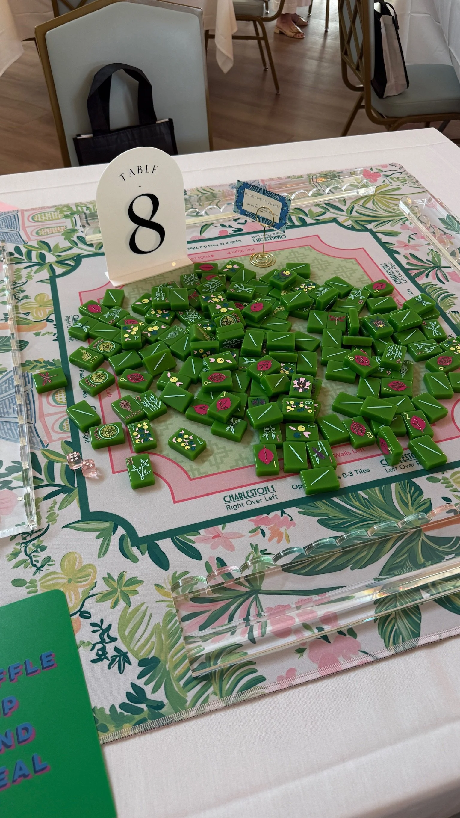 Table with colorful floral design and a game of Mahjong tiles, including a sign indicating Table 8, event schedule and chairs in the background.