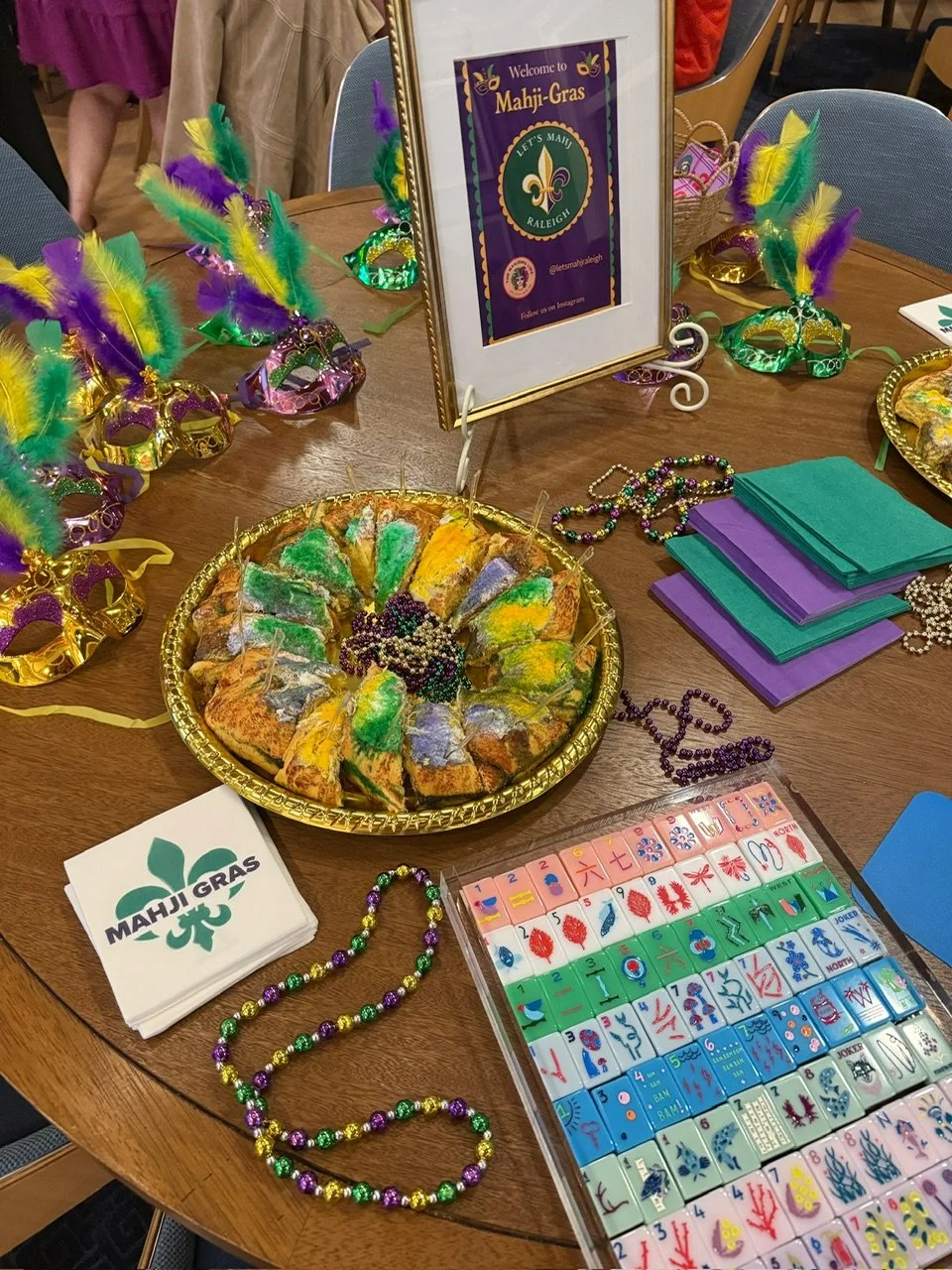 Table decorated for Mahji-Gras with colorful masks, beads, and stacks of purple and green napkins, featuring a plate of colorful, themed cookies, and a sign welcoming to Mahji-Gras.