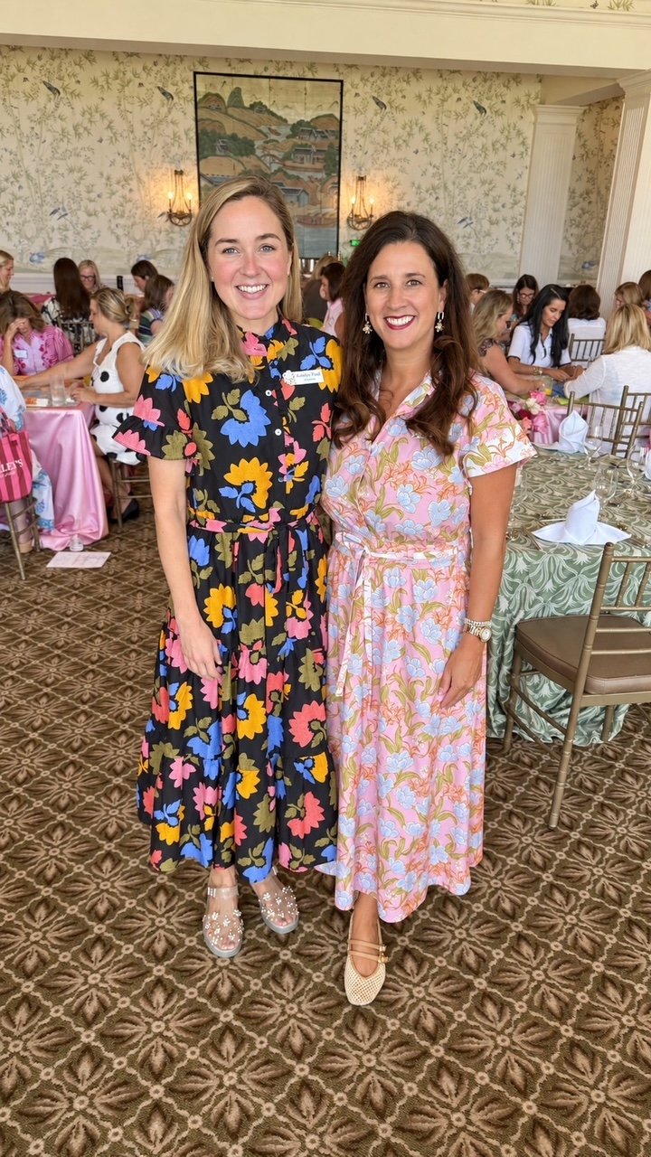 Two women in floral dresses smiling and posing together at a social event with many women at tables in the background.
