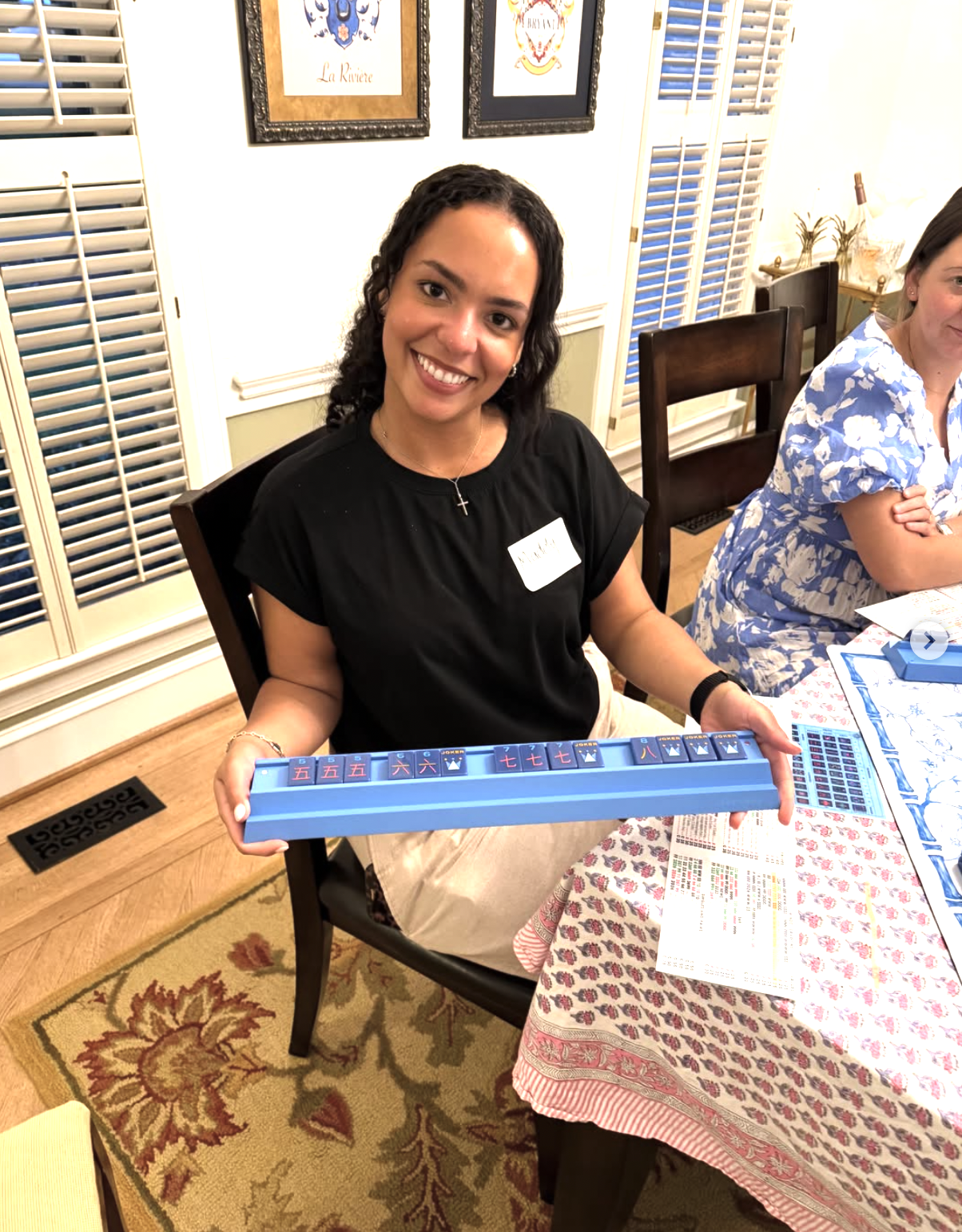 A woman smiling at the camera, sitting at a dining table, holding a blue Mahjong tile set. She has dark curly hair and is wearing a black top with a name tag, a necklace, and a bracelet. The table has a patterned tablecloth with papers on it. Behind her are wooden shutters, framed artwork, and a person partially visible sitting next to her.