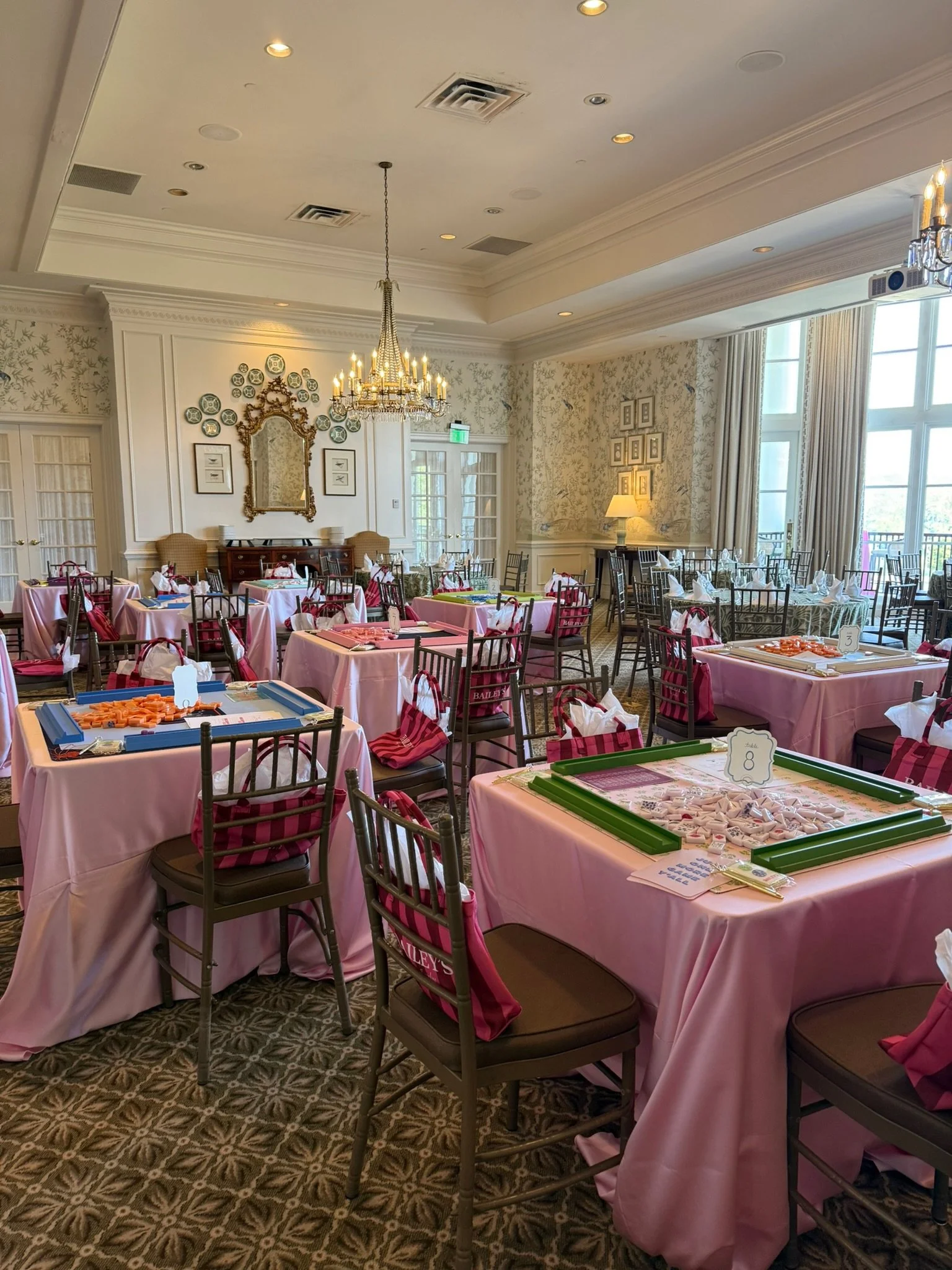 A decorated banquet hall with pink tablecloths, game tables with white tiles, and chairs with pink ribbons. The room features a chandelier, framed pictures, and large windows with curtains, creating an elegant atmosphere.