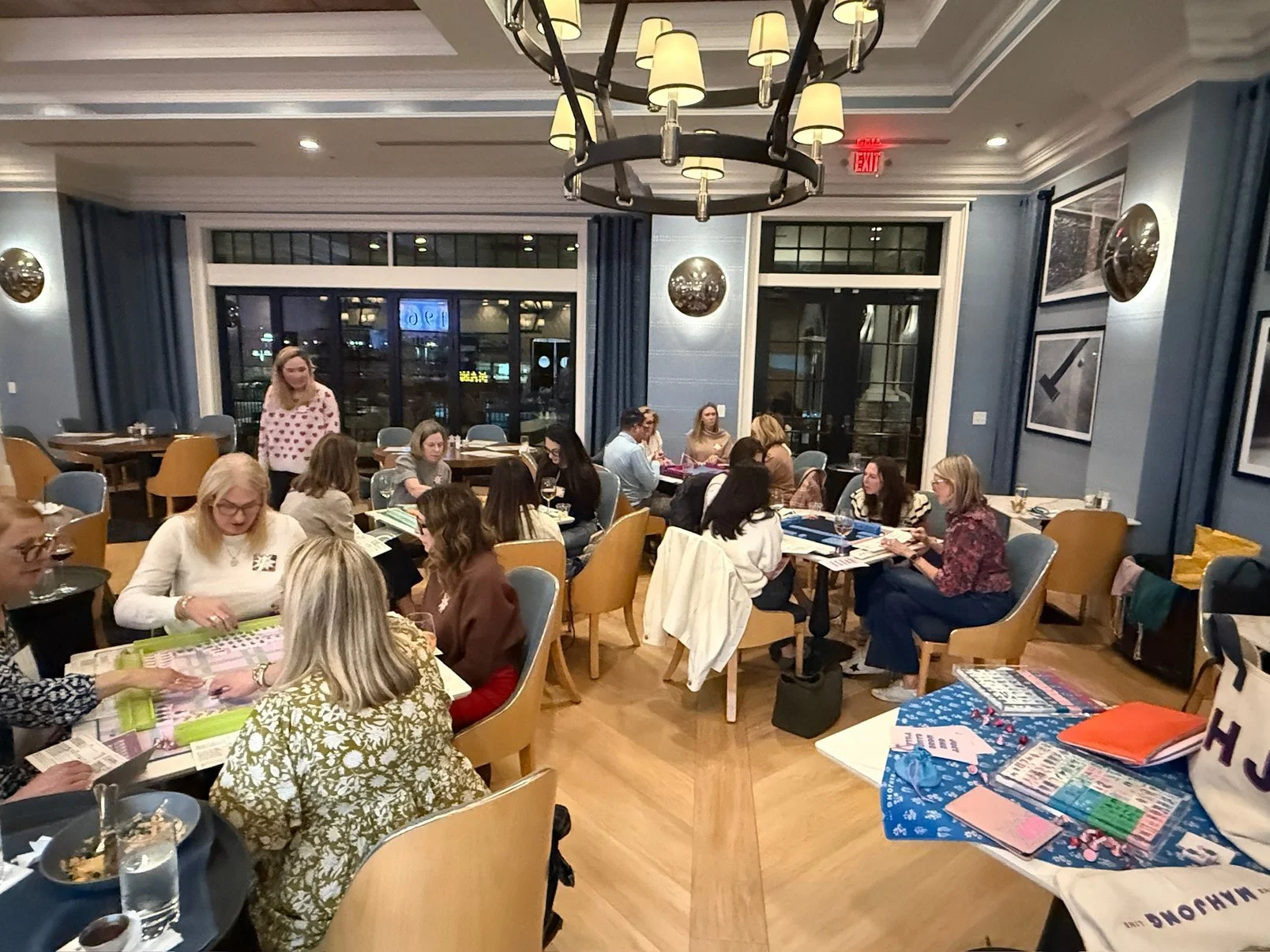 Women gathered in a restaurant, sitting around tables covered with various papers and magazines, engaging in discussion, with some enjoying drinks, in a well-lit, modern space with large windows, blue walls, and framed artwork.