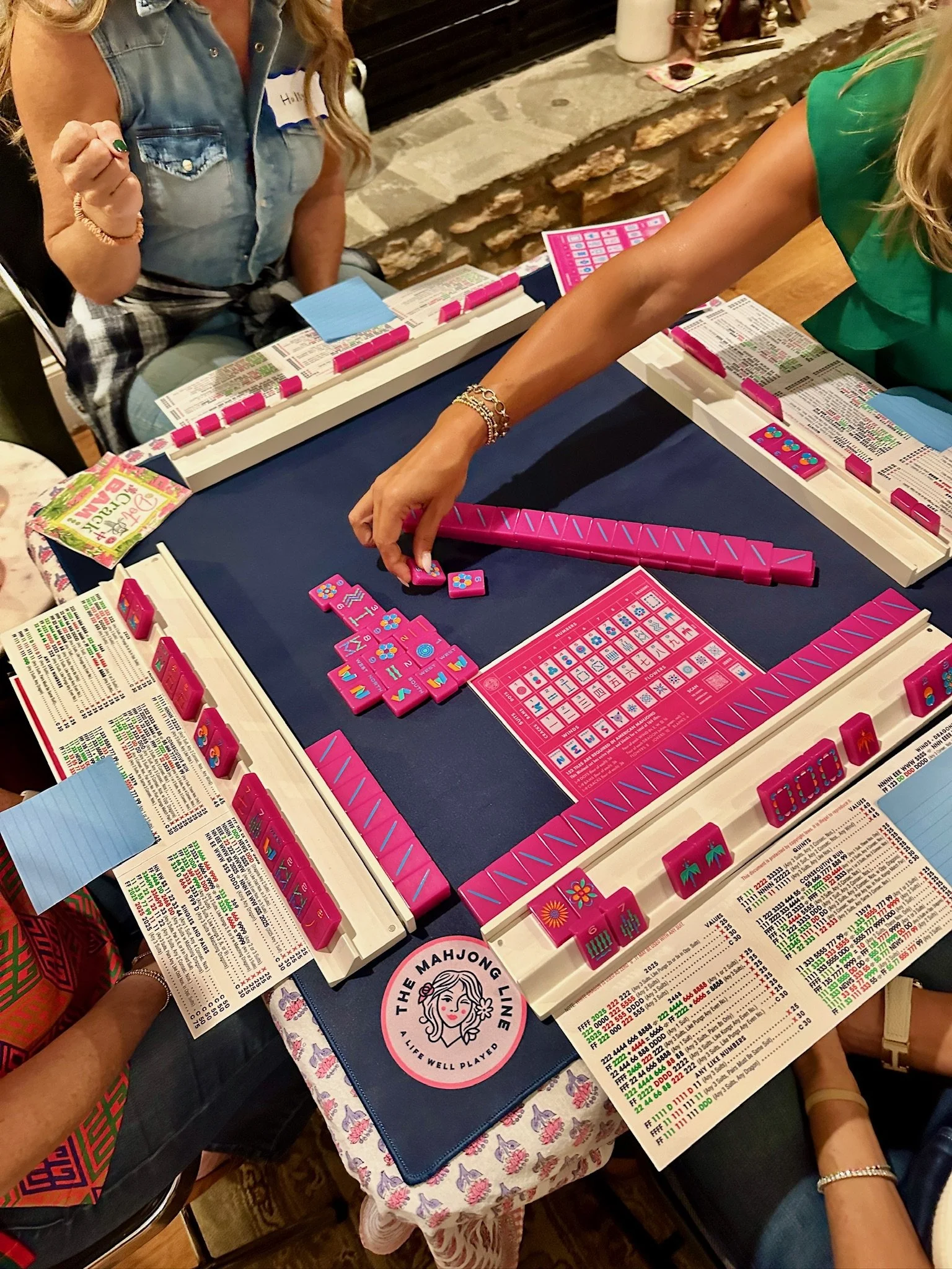 Two women playing Mahjong at a table with Mahjong tiles, score sheets, and drinks in a cozy setting.