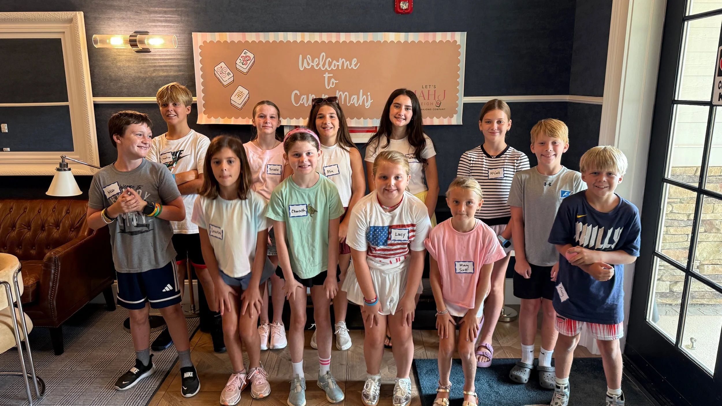 Young children standing together indoors, posing for a photo during a group activity or event. They are smiling, and some are wearing name tags. Behind them is a large sign that says, 'Welcome to Car Mahi,' indicating a gathering or celebration related to mahjong.