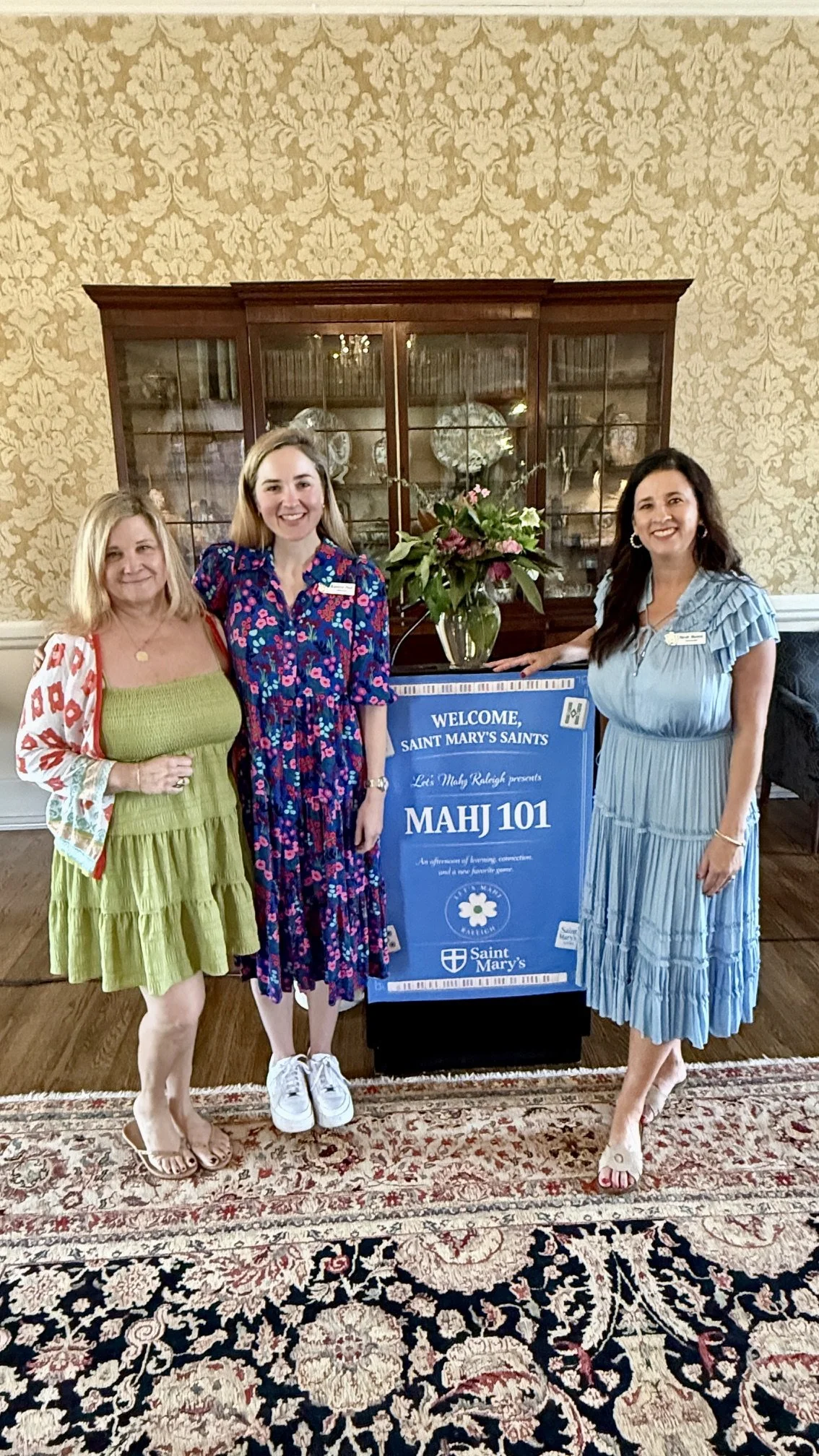 Three women standing in a room with a floral-patterned rug, a wooden china cabinet, and a welcome sign for Saint Mary's Saints with a flower arrangement on top. They are smiling and dressed in colorful, summer dresses.