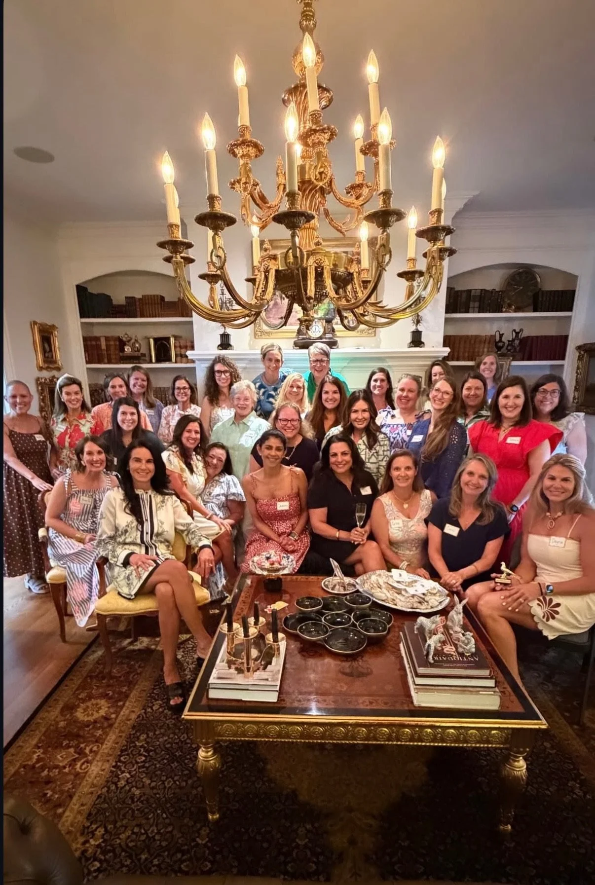 Group of women gathered in a living room, sitting and standing around a coffee table with snacks and drinks, in front of a white fireplace with a large chandelier overhead.