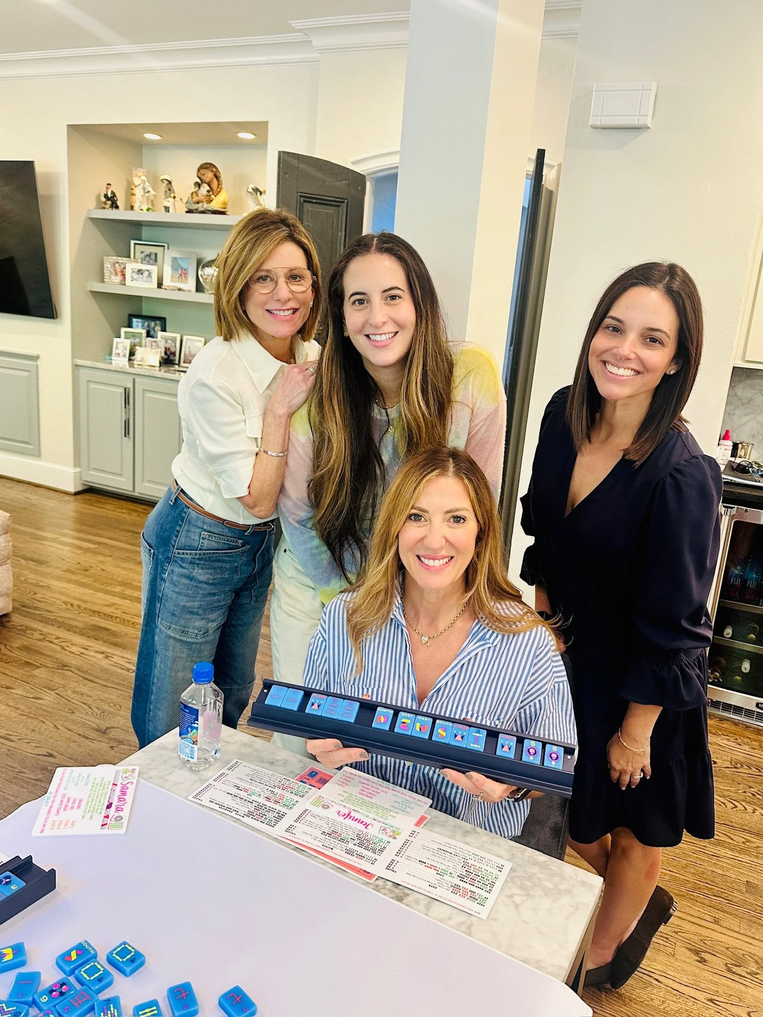 A group of five women gathered around a table at a social gathering, with one woman holding a colorful tile-based game, inside a well-lit room with wooden floors and white walls.