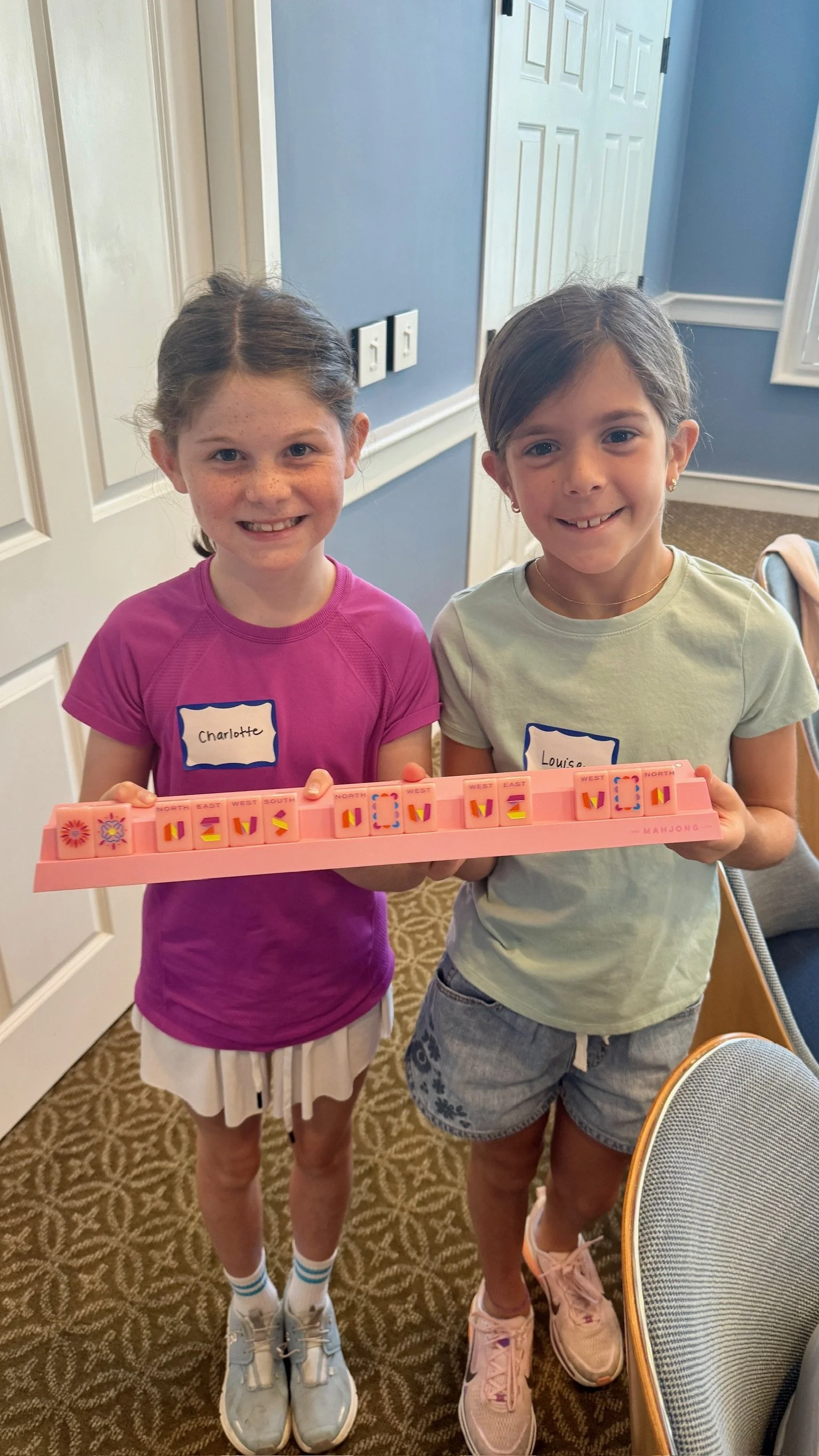 Two young girls standing indoors, smiling, holding a pink Mahjong tile rack. The girl on the left is wearing a pink shirt and white shorts, while the girl on the right is wearing a light green shirt and denim shorts. The girl on the left has a name tag that says 'Charlotte', and the girl on the right has a name tag that says 'Louise'.